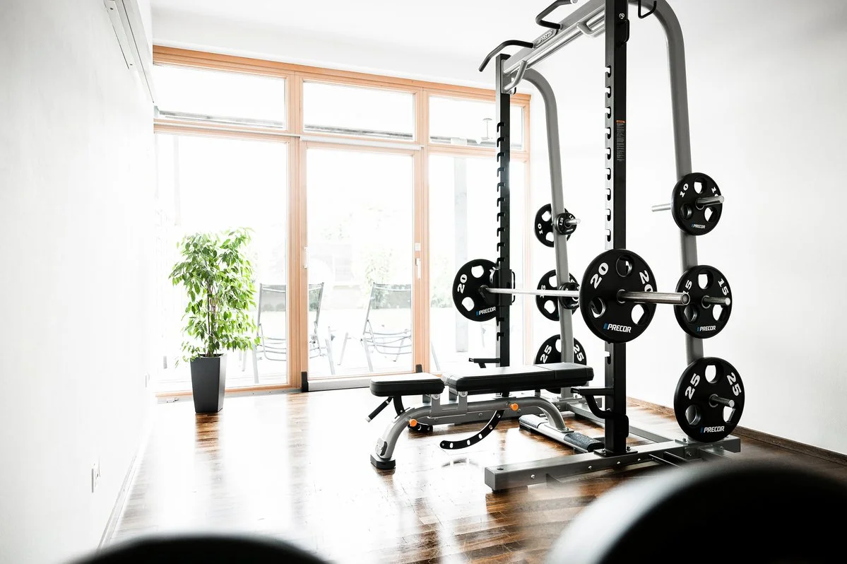 Home gym with weightlifting equipment in a bright room with large windows, hardwood floors, and a potted plant.