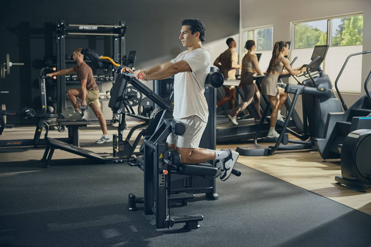 People exercising in a gym on various cardio and strength machines, with natural light coming through large windows.