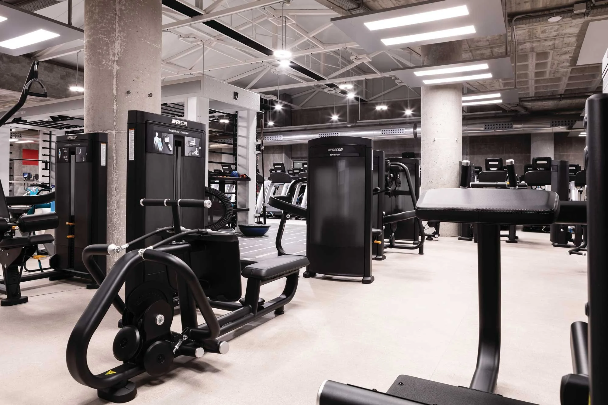 Empty gym with various workout equipment, including weight machines, treadmills, and stationary bikes, in a modern industrial-style space with concrete pillars and ceiling.