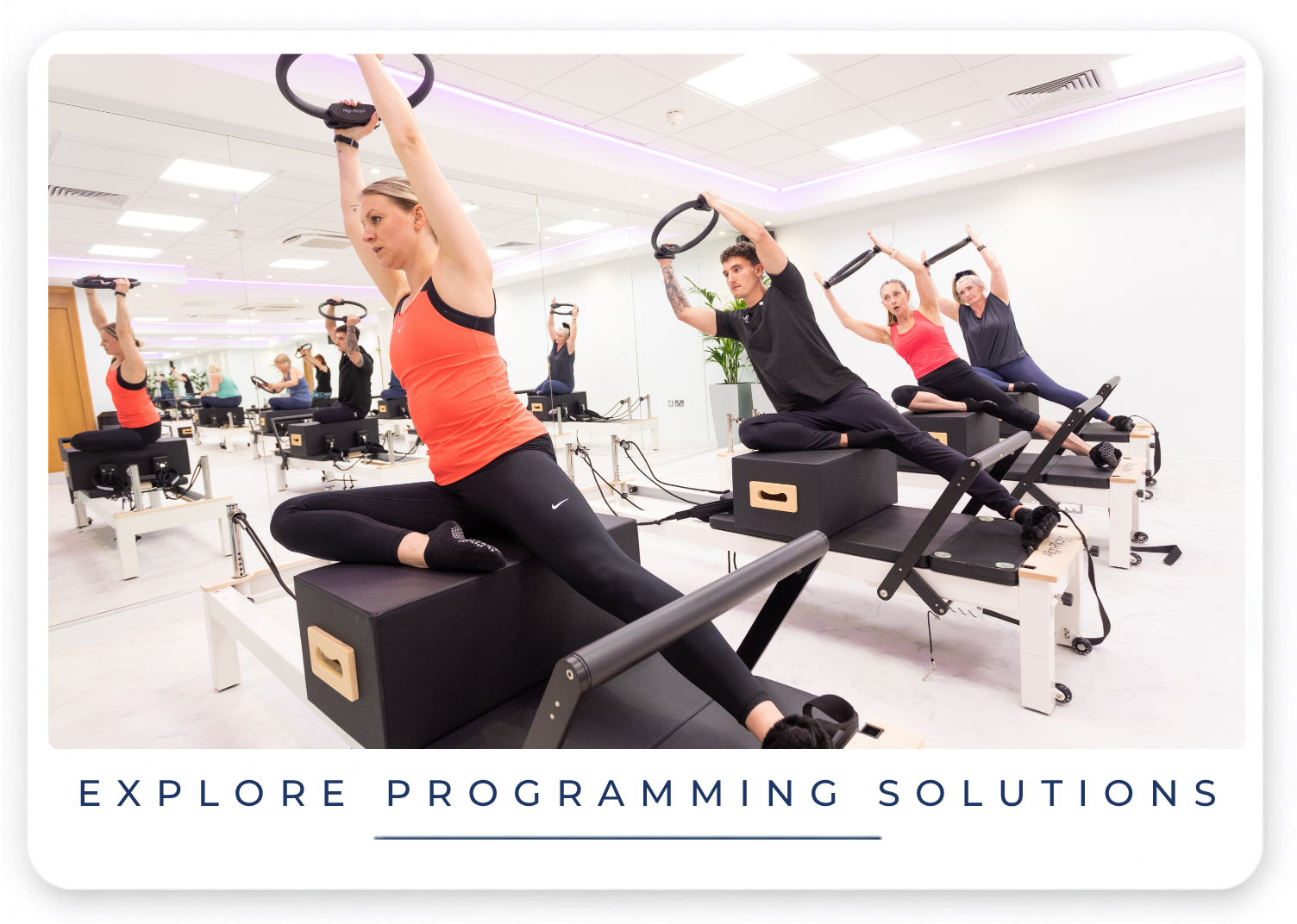 A group of people participating in a Pilates or fitness class using specialized equipment that involves rings. The room has mirrored walls and soft lighting, with a clean, modern design and a white floor.