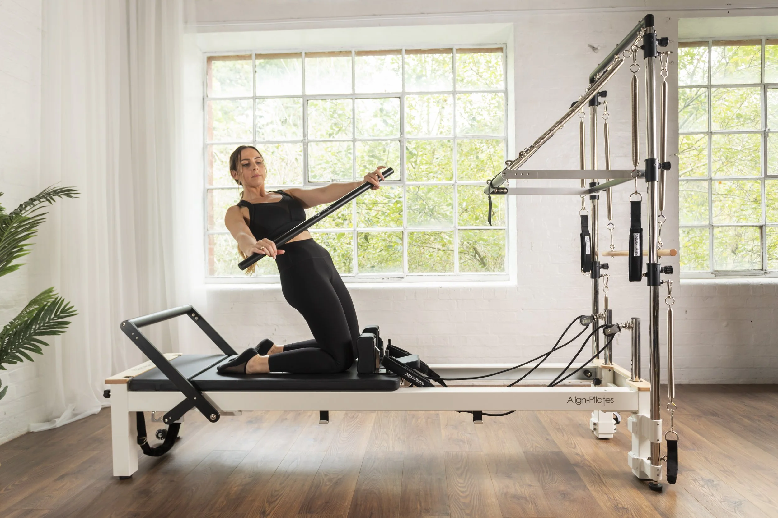 Woman in black workout attire exercising on a Pilates reformer machine in a bright room with large windows and wooden floors.