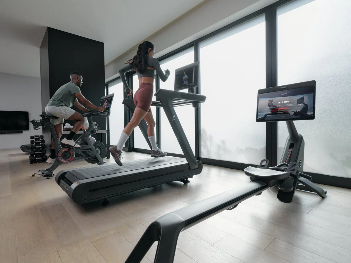 People exercising on a treadmill and stationary bike in a modern gym with large windows and a TV.