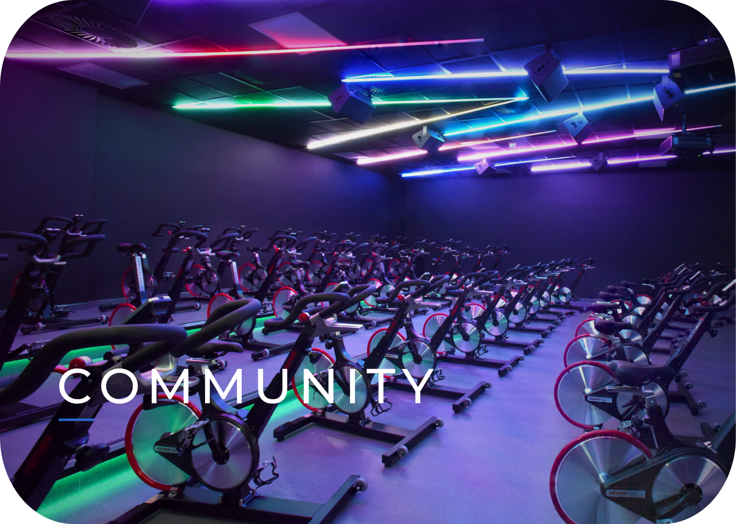 A gym filled with rows of stationary exercise bikes illuminated by colorful neon lights on the ceiling, creating a vibrant atmosphere. The word 'COMMUNITY' is overlaid on the lower part of the image.