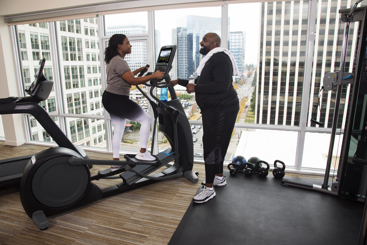 A woman running on a treadmill in a gym, conversing with a man holding a water bottle, with large windows showing a cityscape in the background.