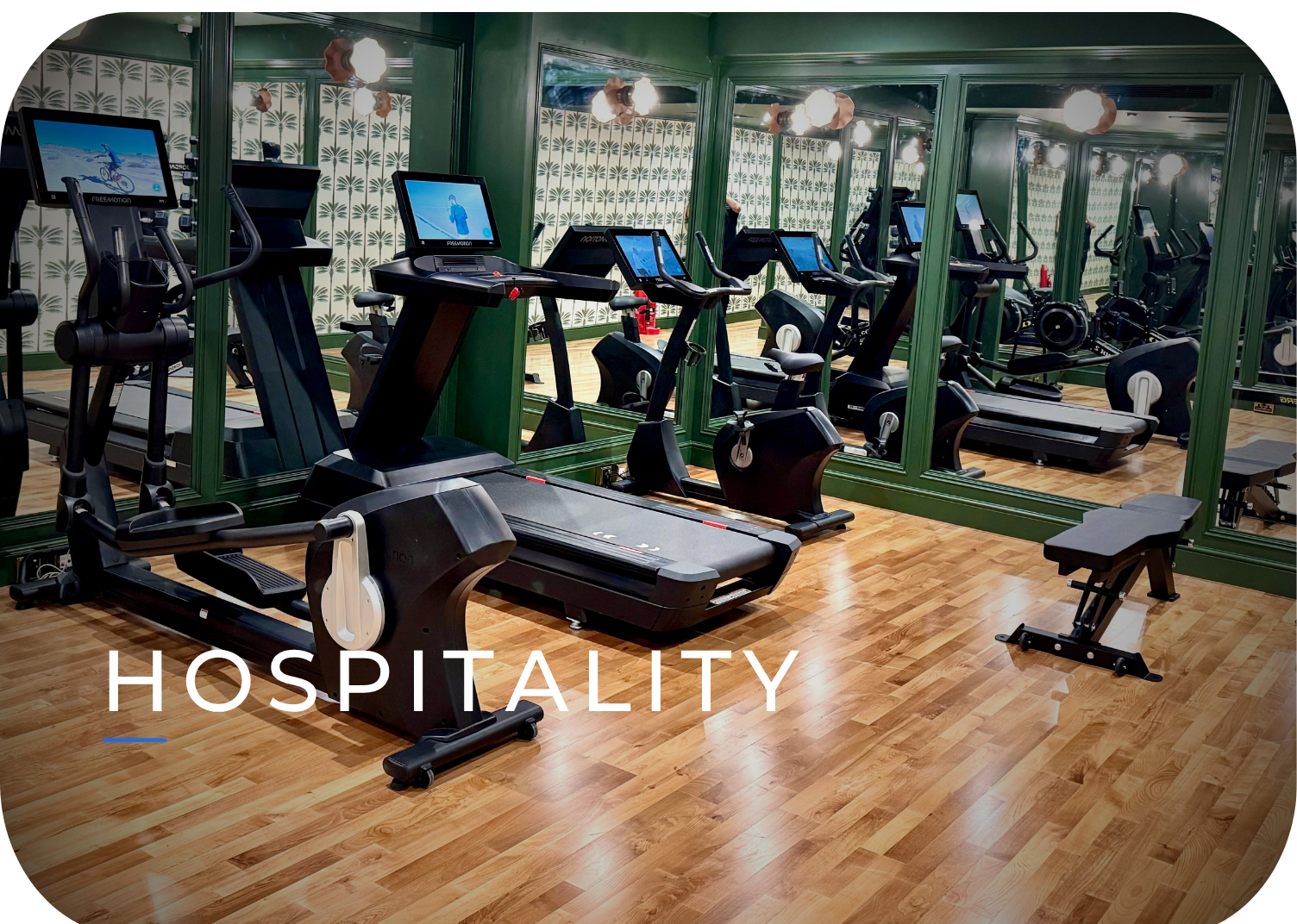 Empty gym with various cardio equipment including treadmills, ellipticals, and stationary bikes, with mirrored walls and wooden floors, and the word 'HOSPITALITY' overlaid.
