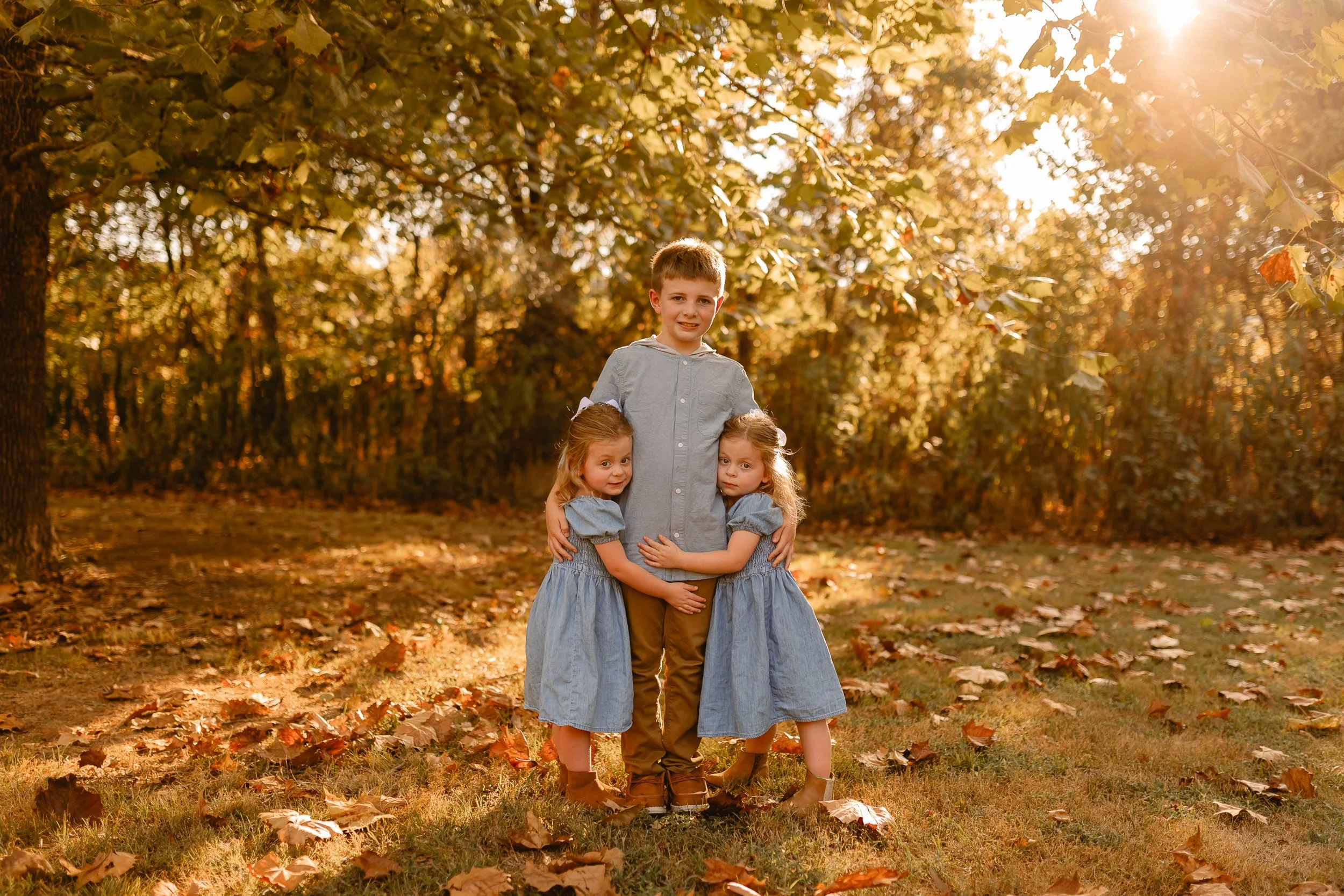 A boy is standing in the center next to two young girls, all hugging each other outdoors on a fall day. The scene is illuminated with warm sunlight, and fallen leaves cover the ground amid trees with yellow and orange leaves.