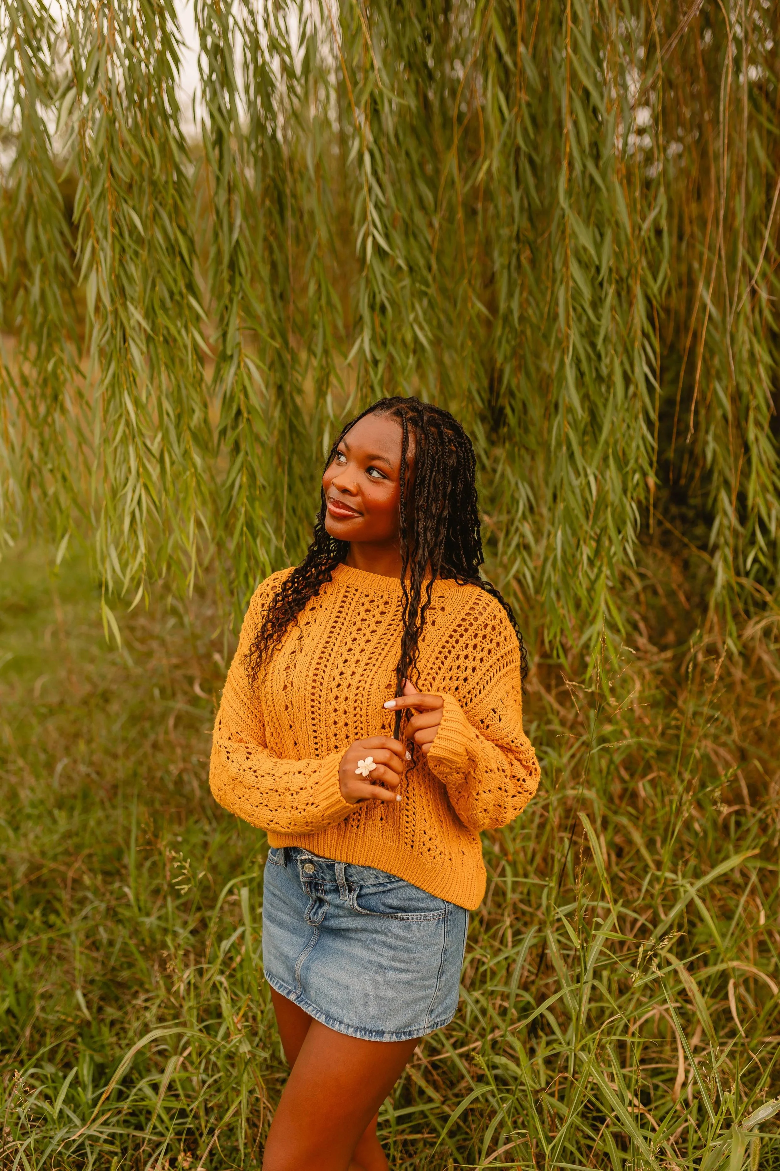 A woman with long braided hair standing in front of a large weeping willow tree, wearing a mustard yellow knit sweater and a denim skirt, with a large white flower ring on her finger.