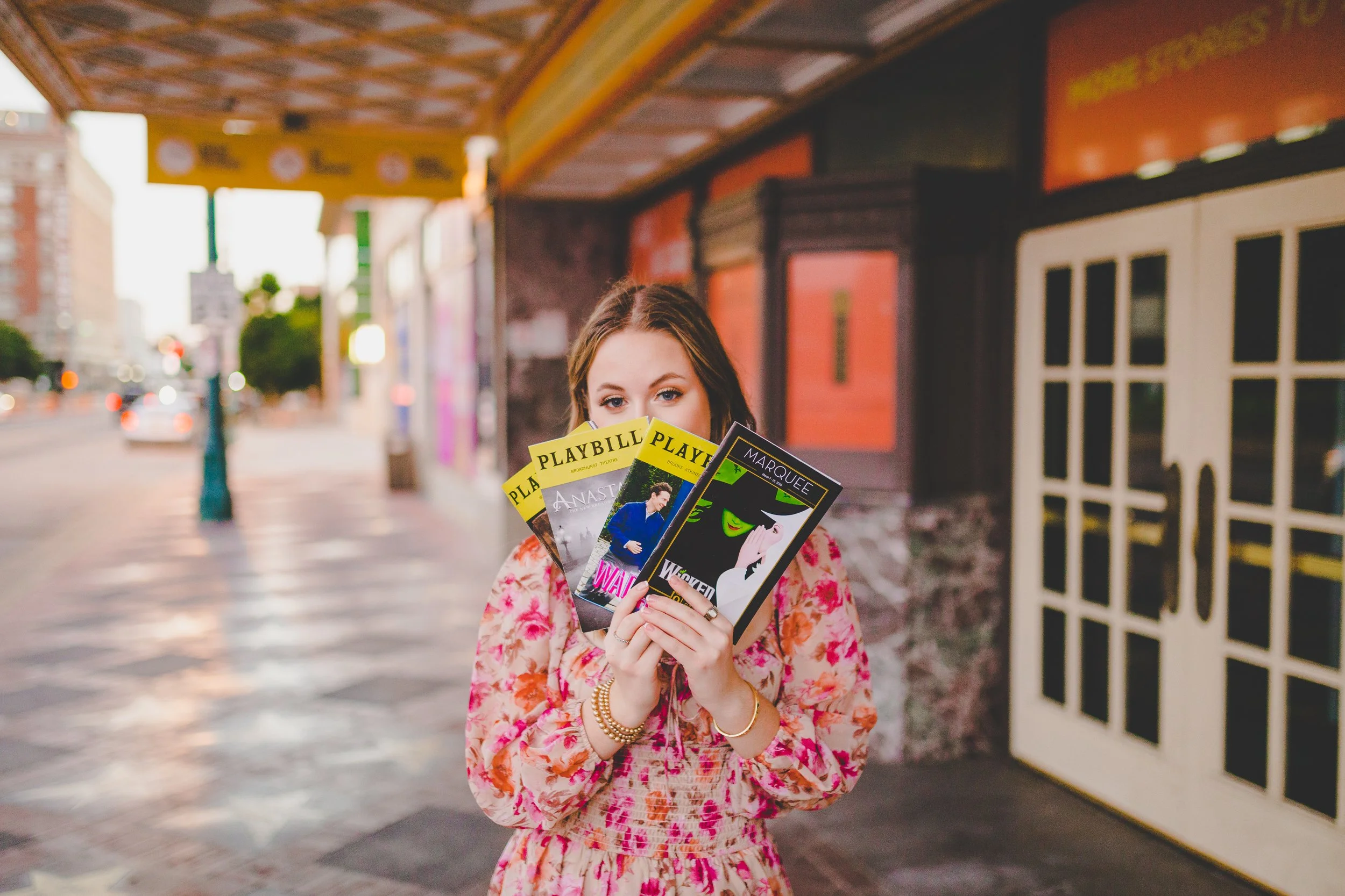 A young woman with brown hair holding several theater playbills partially covering her face, standing outdoors on a city sidewalk with buildings and traffic in the background during daytime.