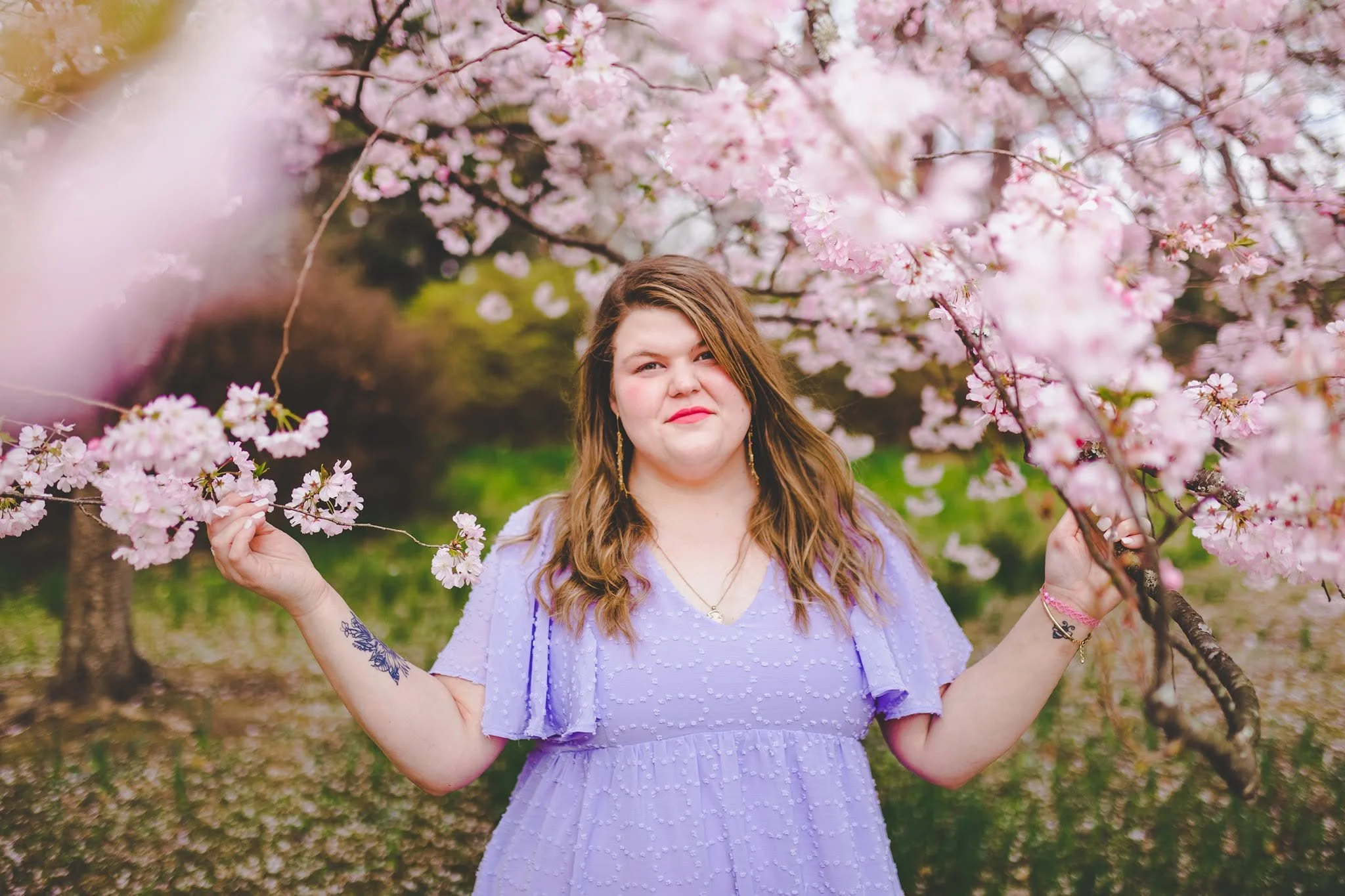 A young woman with brown hair and earrings standing amidst pink cherry blossom trees, holding branches with blossoms.