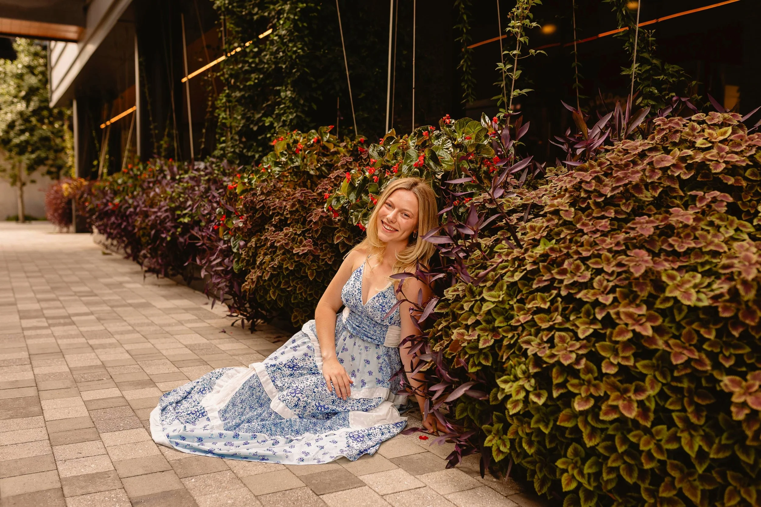 A woman sitting on the sidewalk next to a large hedge of purple and green plants with red flowers, smiling and wearing a blue and white patterned dress.