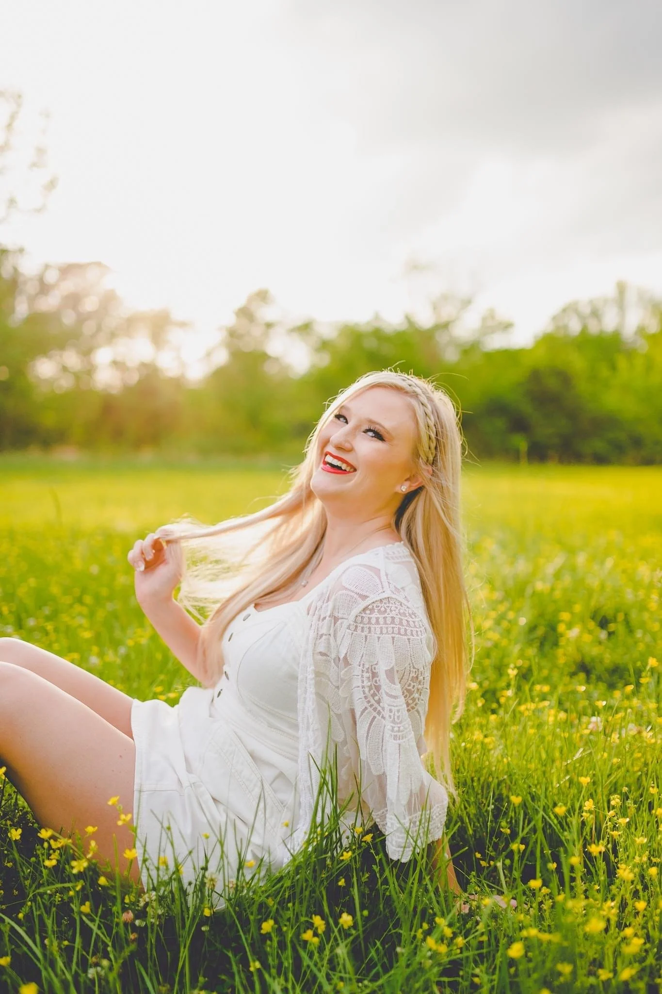 Young woman with long blonde hair and a white lace dress sitting in a field of yellow flowers, smiling and looking up, during daytime with overcast sky.