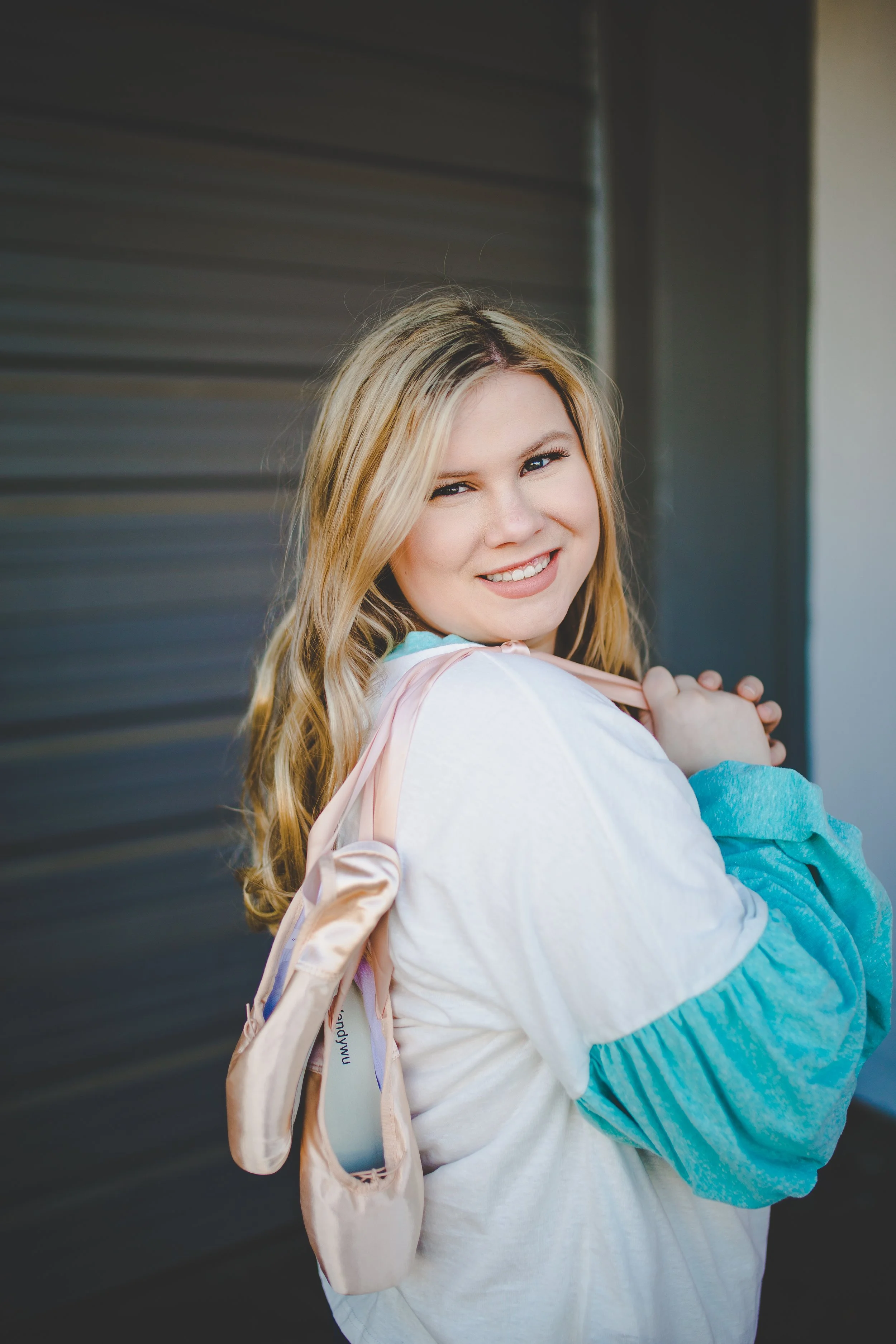 A young woman with long blonde hair smiling, wearing a white shirt and a turquoise jacket, carrying a light pink bag on her shoulder, standing outdoors against a dark, textured background.