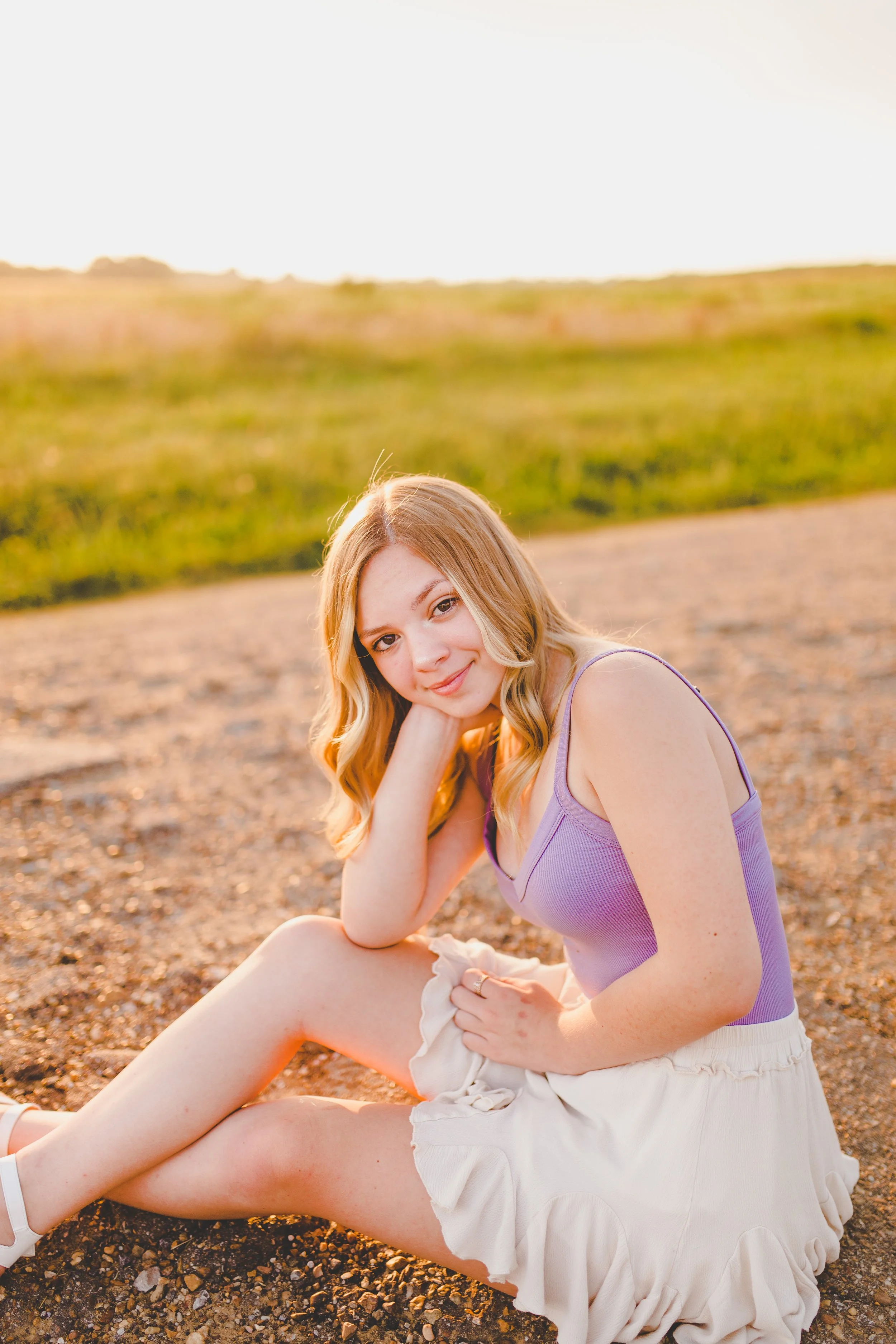 A young woman with long blonde hair, wearing a lavender sleeveless top and a white skirt, sitting on a gravelly ground outdoors during sunset, smiling at the camera with a pull-back view of a landscape with grass and hills in the background.