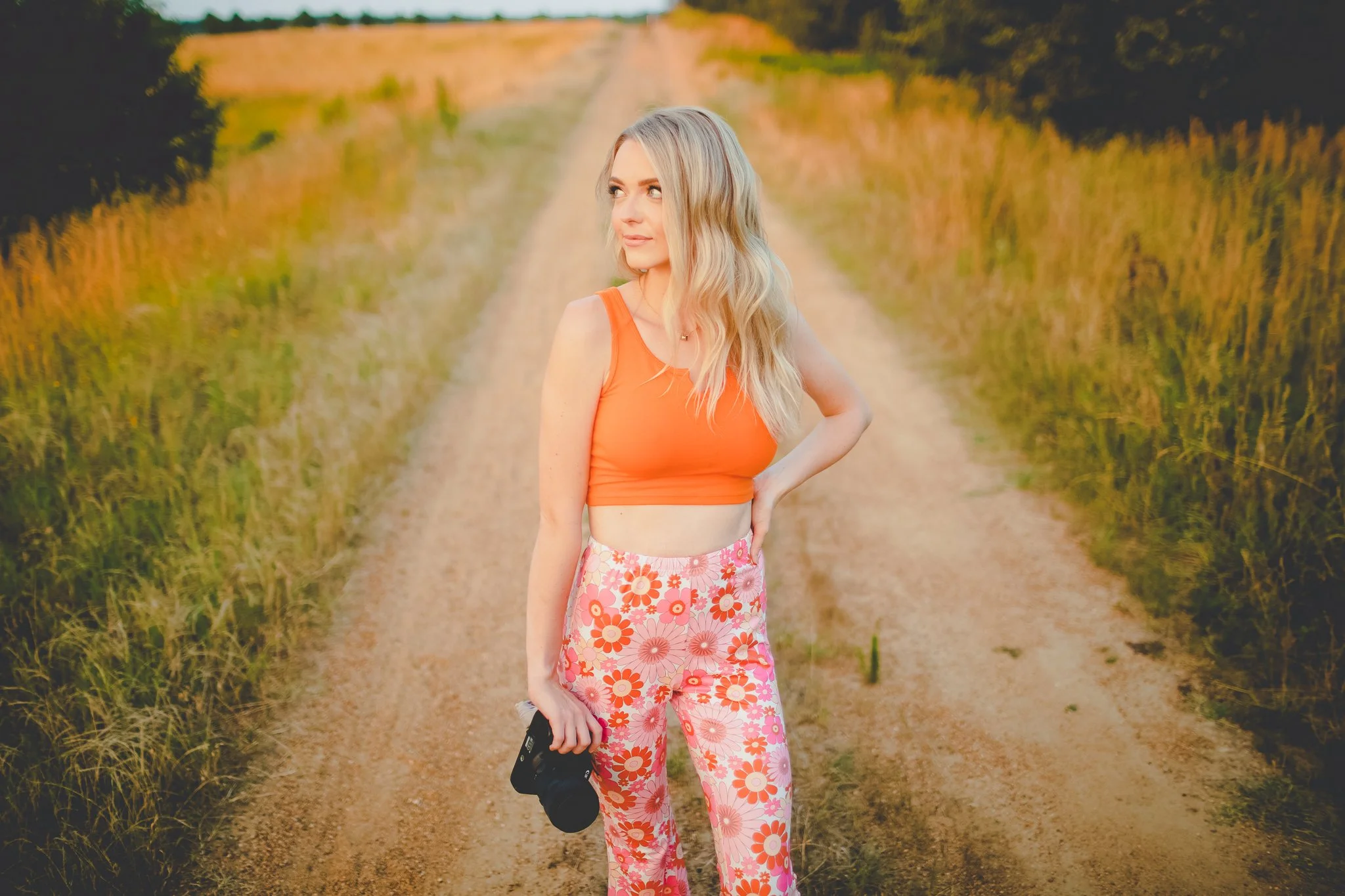 A young woman stands on a dirt road in a rural area, holding a camera in her right hand, wearing a bright orange top and pink patterned pants, with blonde wavy hair, during late afternoon or early evening.