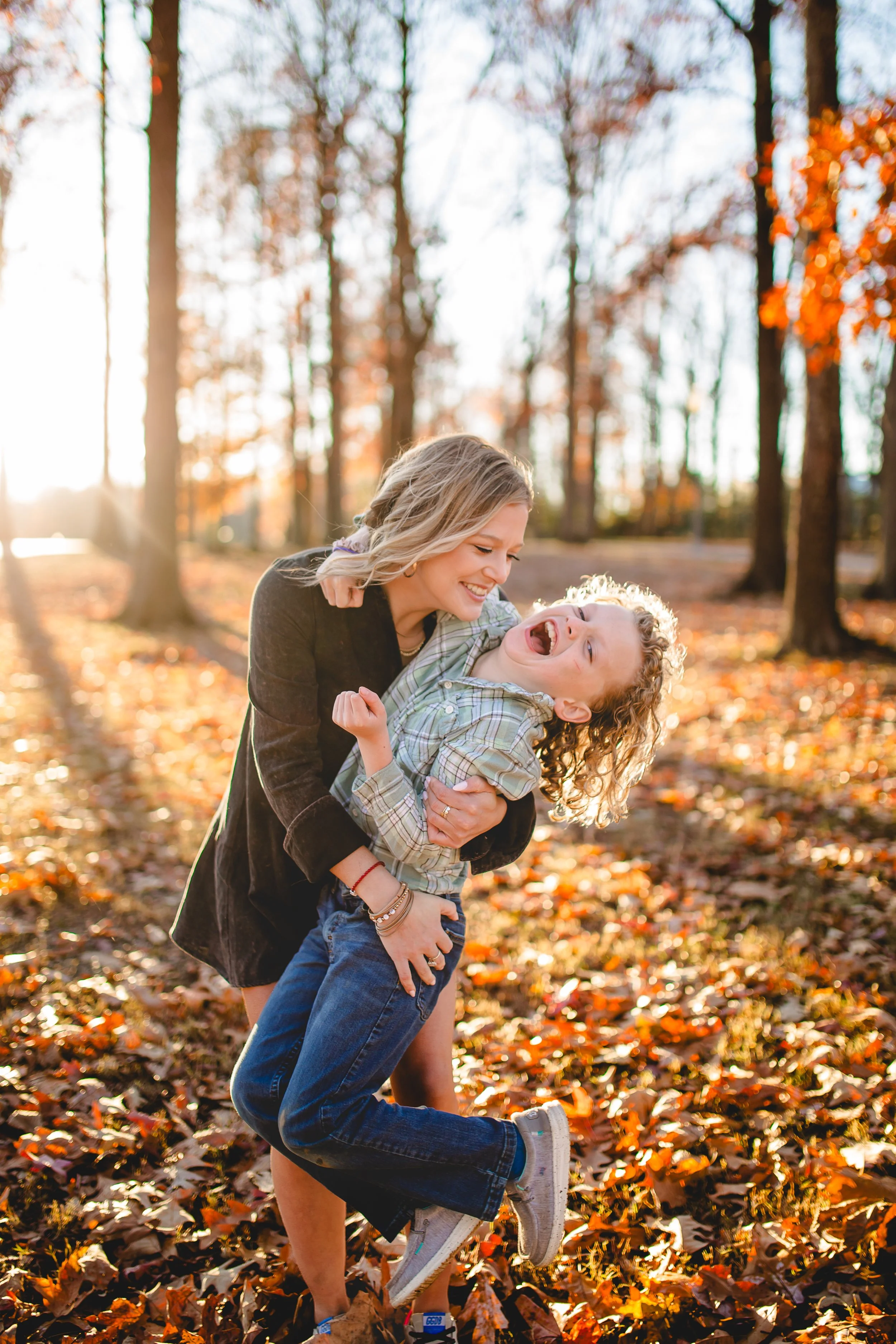 A young woman with blonde hair playing with a young boy with curly hair in a park covered with fallen autumn leaves. They are smiling and having fun during sunset.