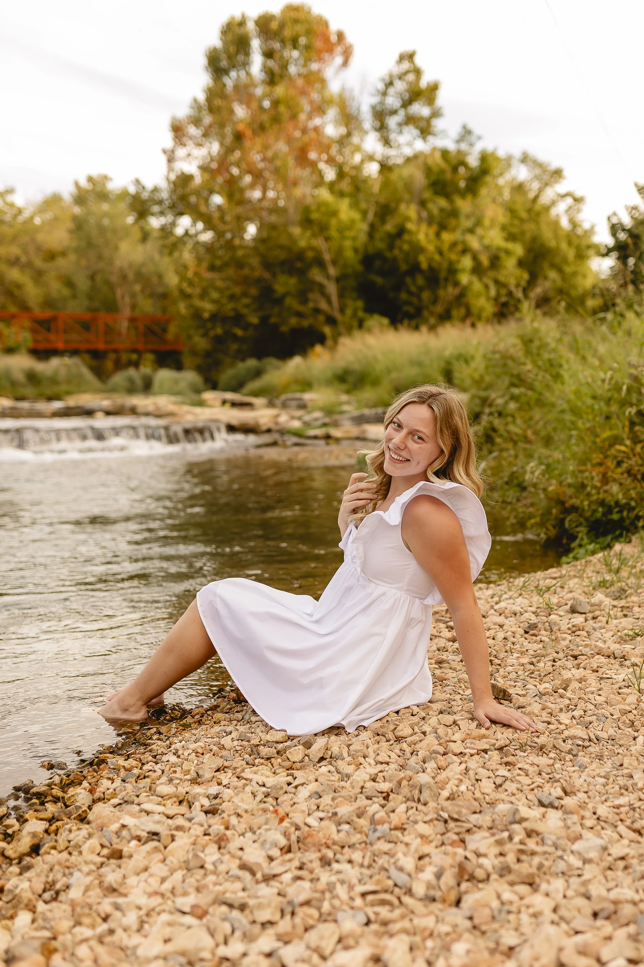 A woman with blonde hair in a white dress sitting on the rocky shore of a river, smiling and leaning back with her feet in the water. In the background, there are trees with autumn leaves and a small bridge over the river.