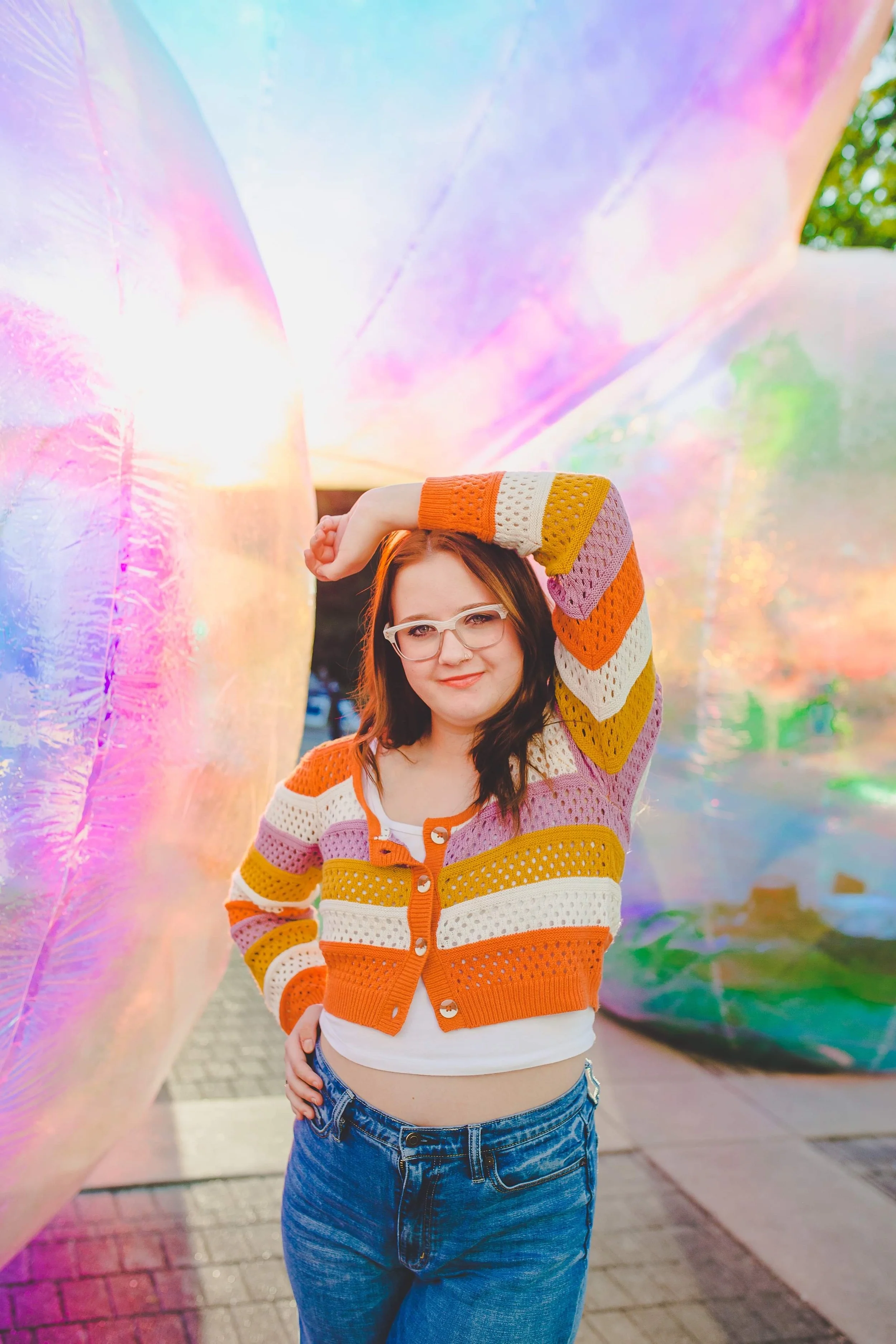 A woman with glasses standing outdoors posing with one hand raised above her head, wearing a colorful striped sweater and blue jeans, behind a large iridescent balloon or balloon wall.