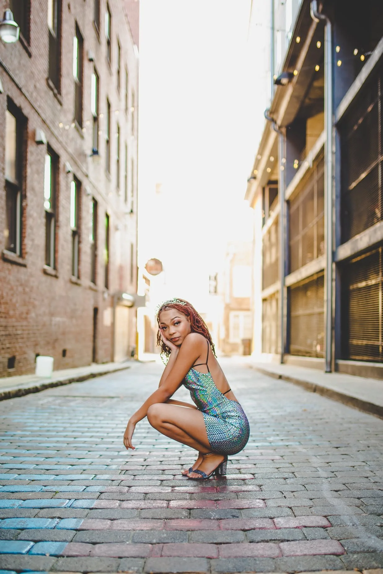 A woman with red hair wearing a shiny, patterned dress crouches on a cobblestone street in an alleyway between brick and metal buildings, with bright sunlight behind her.