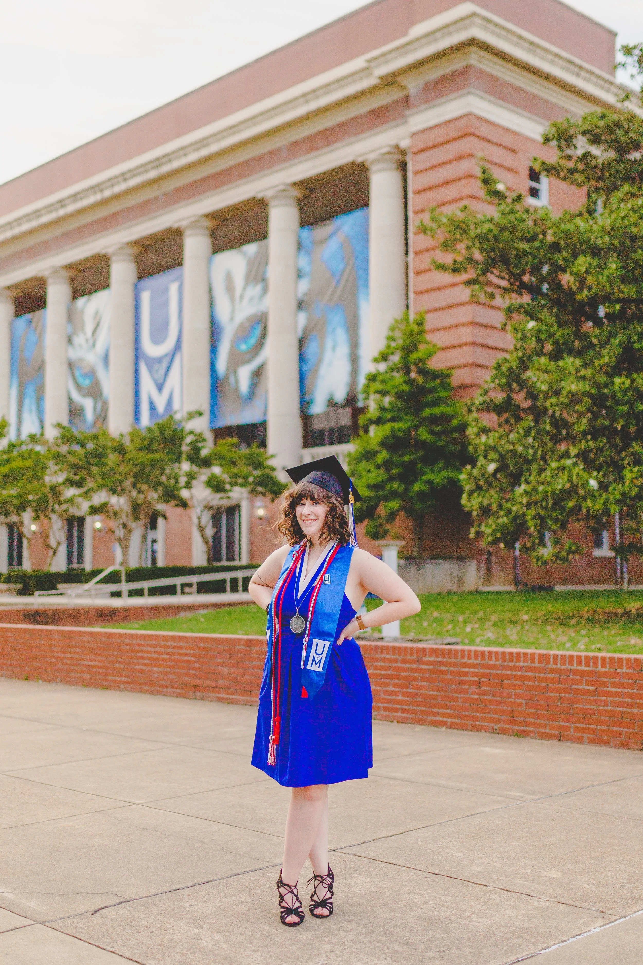 A young woman in a blue graduation gown and cap, wearing medals and ribbons, standing outdoors in front of a brick and stone academic building with large columns and a mural with the letters 'U' and 'M' in the background. She is smiling and posing co