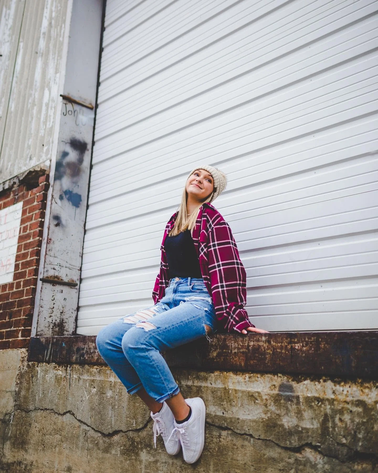 A young woman with long blonde hair wearing a beige knit beanie, black t-shirt, red plaid shirt, ripped jeans, and white sneakers, sitting on a concrete ledge in front of a white corrugated metal wall, smiling and looking up.