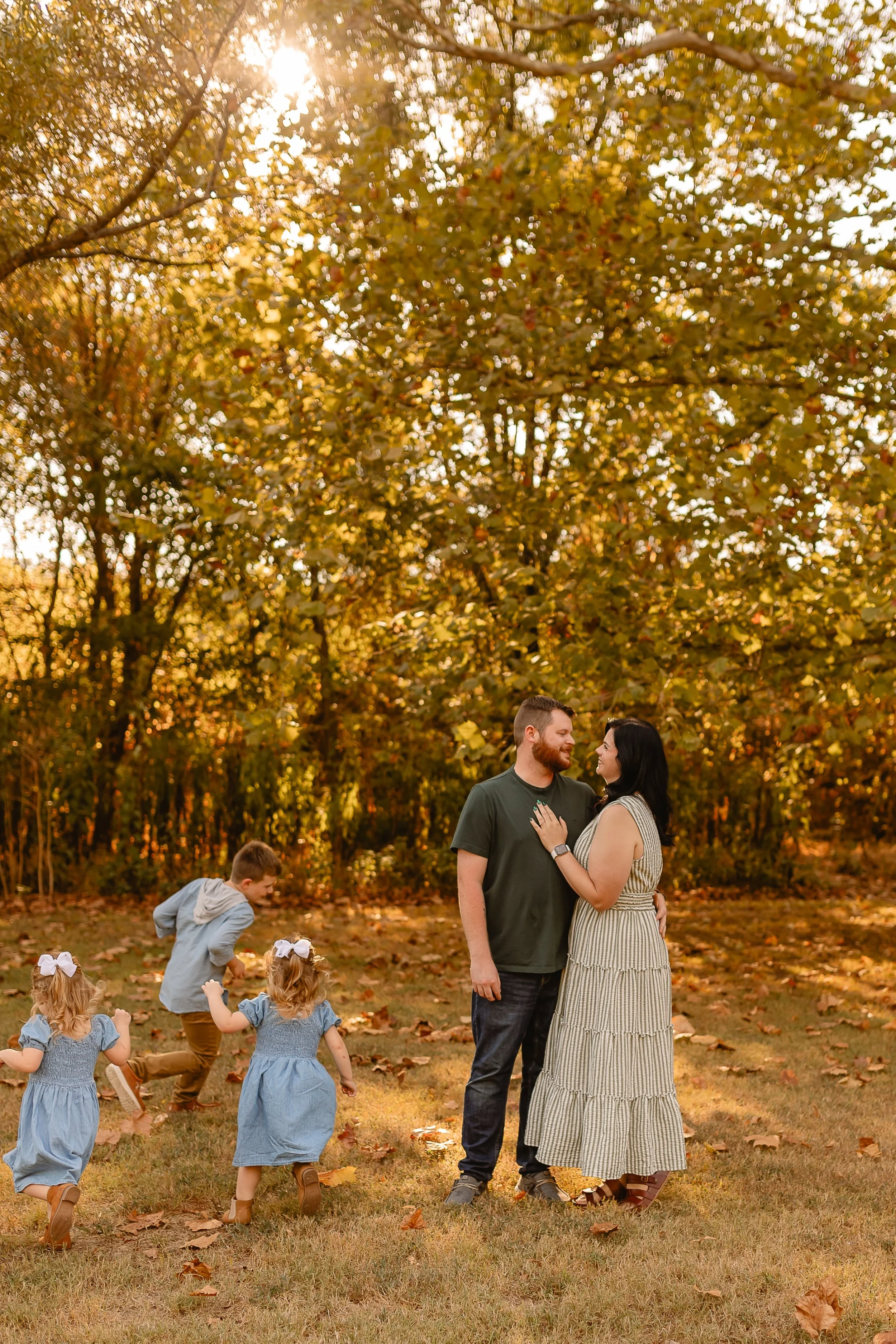 A couple standing close together outdoors during autumn, with children playing nearby in a park surrounded by trees with fall foliage, sunlight filtering through the leaves.