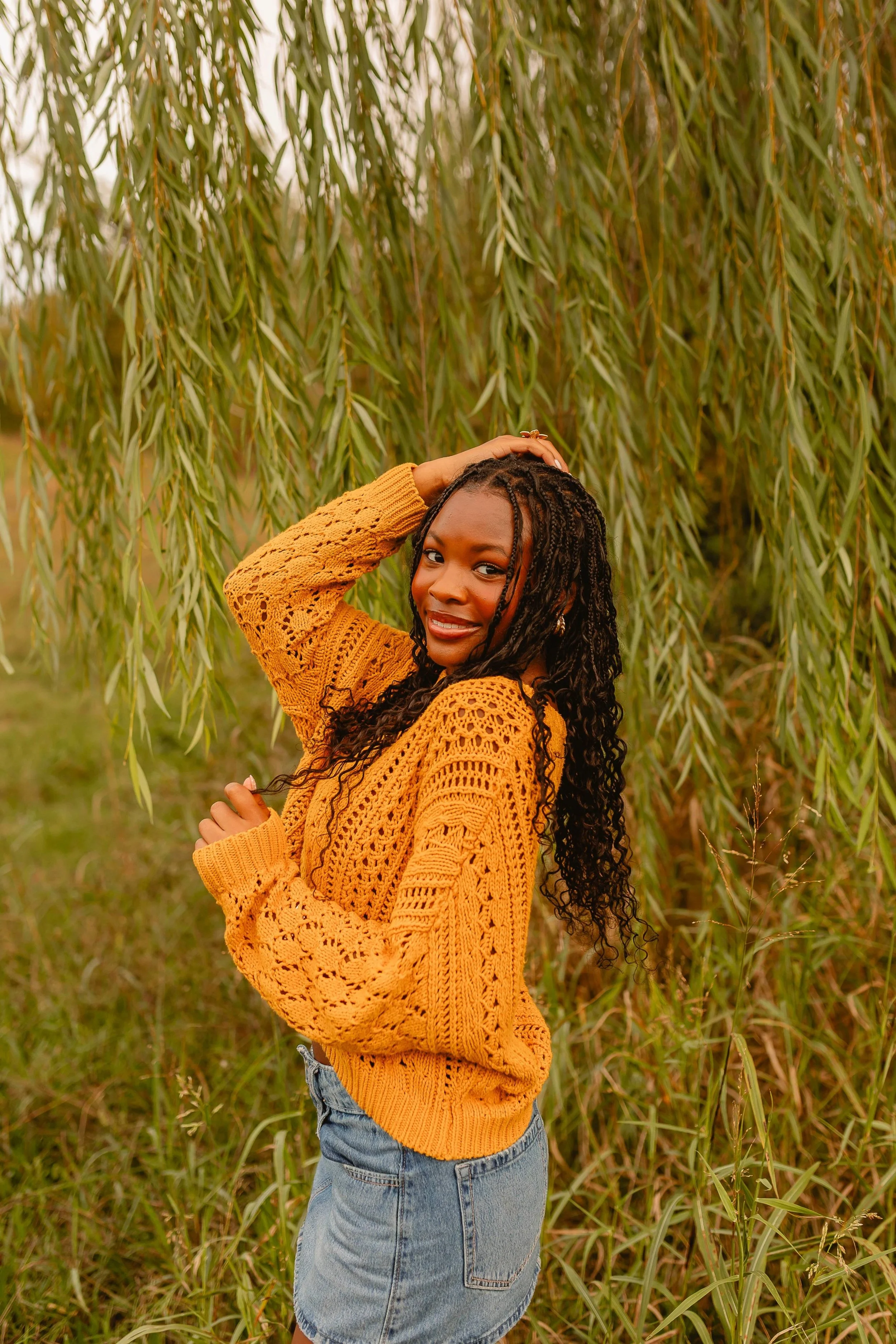 A woman wearing a yellow knitted sweater and a denim skirt poses amid tall grass and a leafy tree, smiling and touching her hair with one hand.