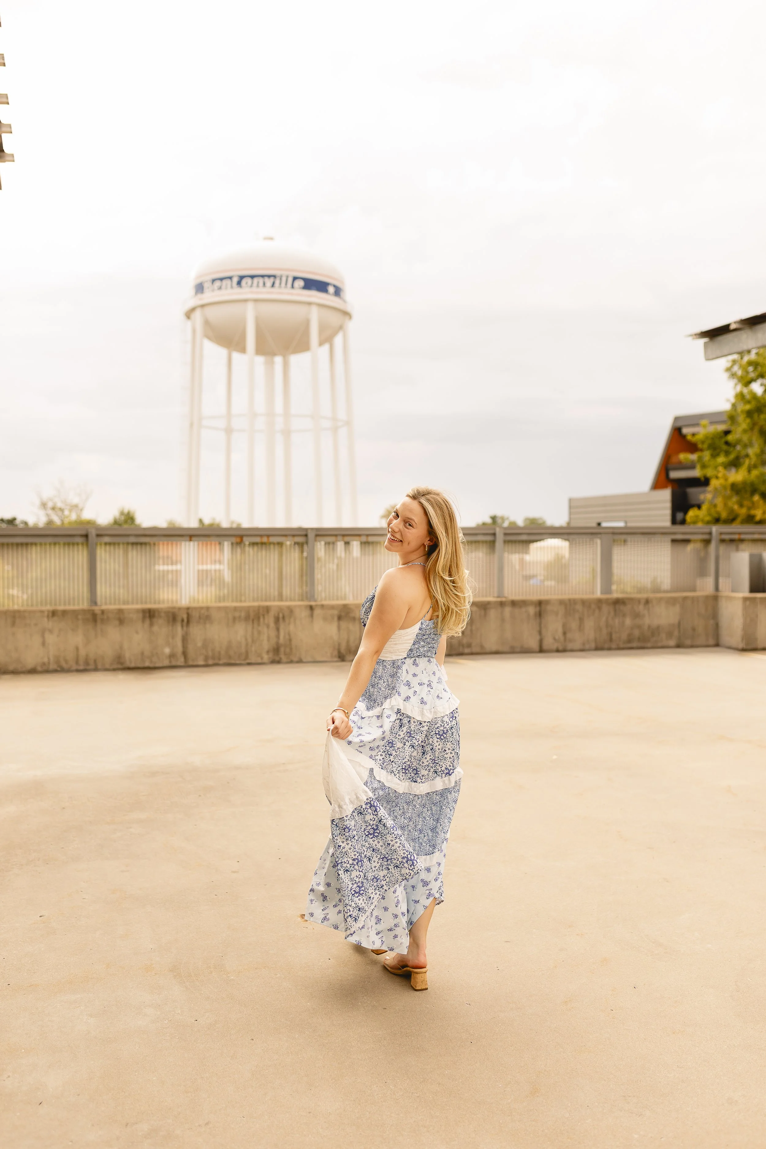A woman in a blue and white floral dress walking barefoot on a rooftop or terrace, smiling, with a water tower and a house in the background.