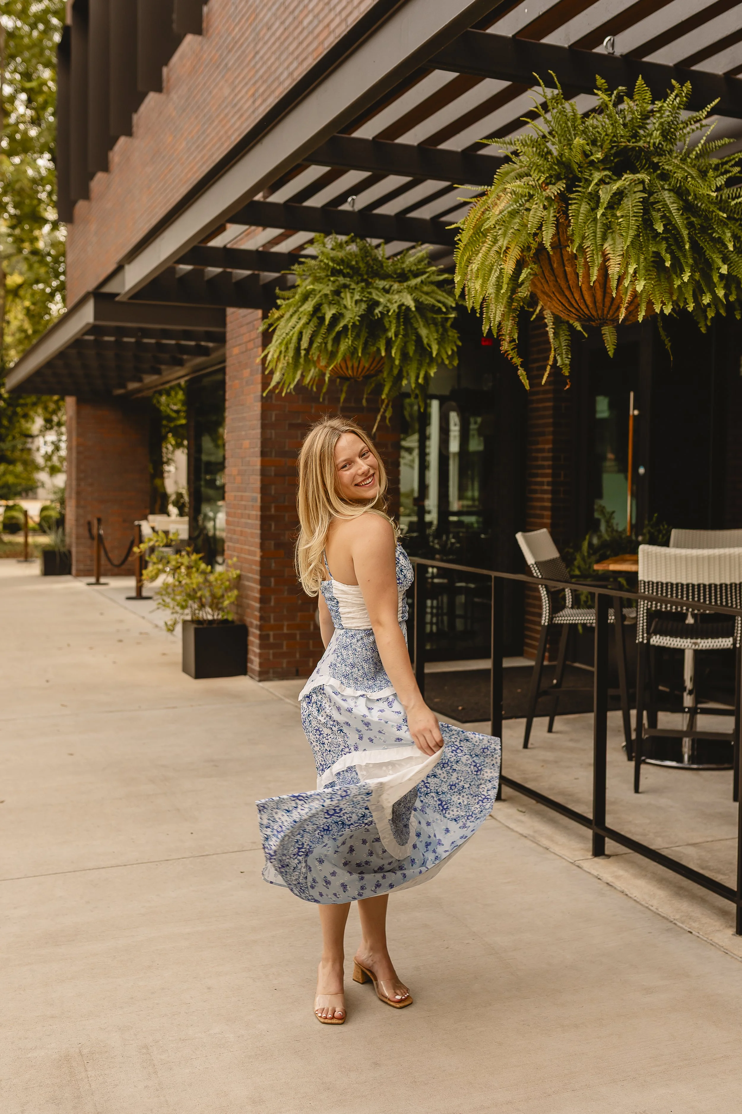 A woman with long blonde hair wearing a blue and white floral dress and tan open-toe heels, dancing outdoors on a sidewalk, with a brick building and hanging plants in the background.