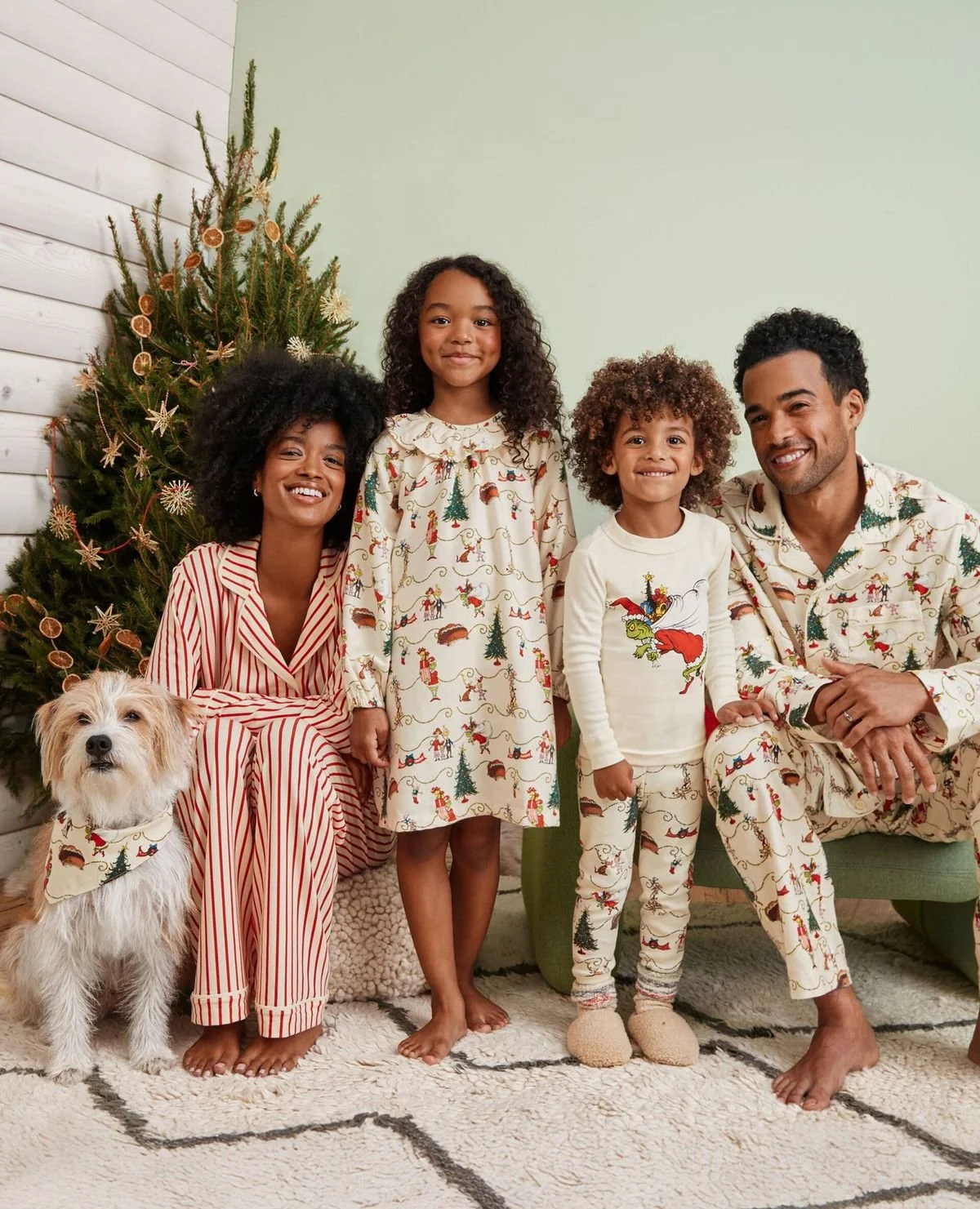 A family of four, including two children, a woman, and a man, along with a dog, poses near a decorated Christmas tree. They are all dressed in festive pajamas with Christmas-themed designs, standing on a rug in a cozy room with a light green wall.