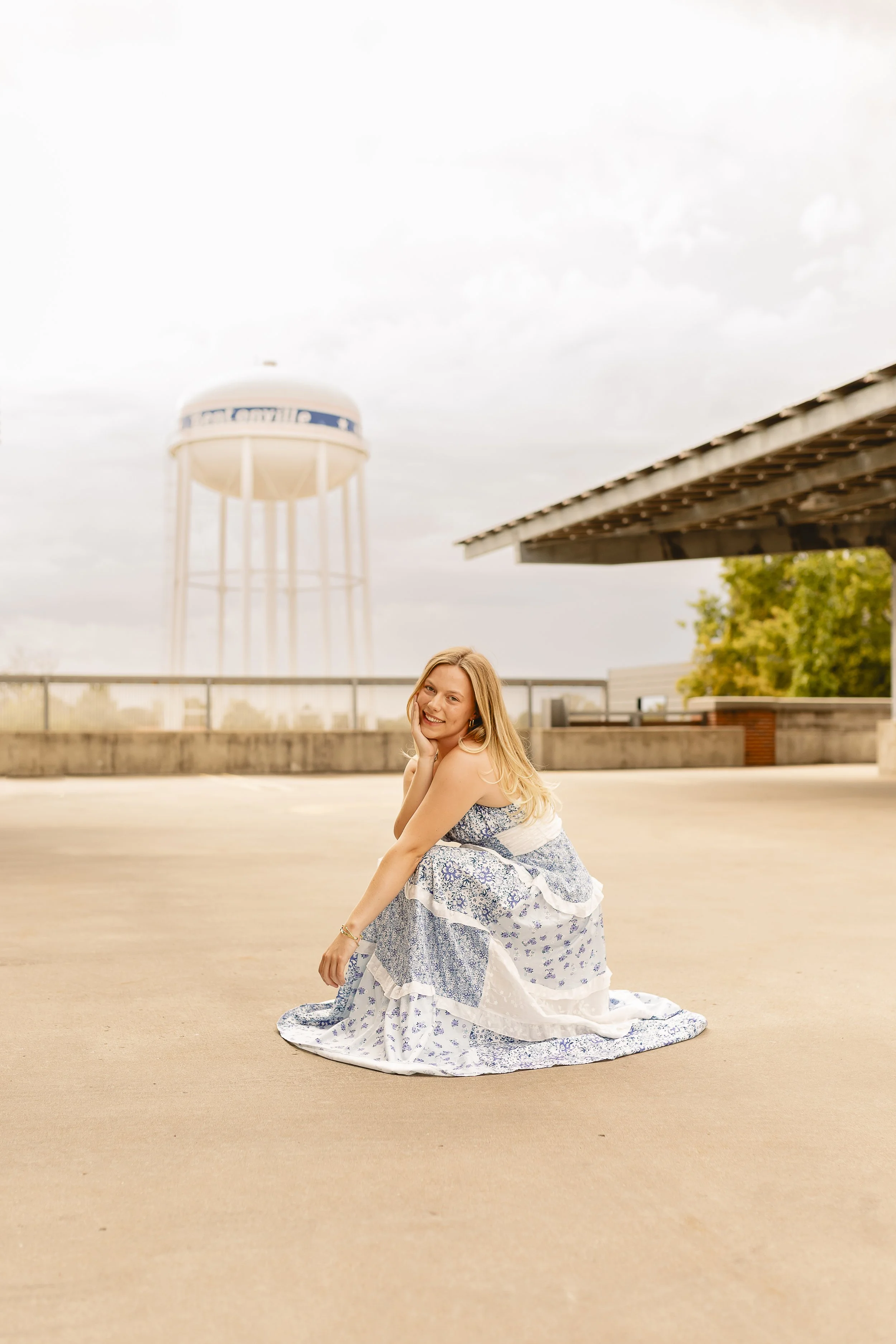 A young woman with long blonde hair sitting on the ground in a white and blue floral dress, smiling and resting her chin on her hand outdoors on a cloudy day, with a water tower and building in the background.