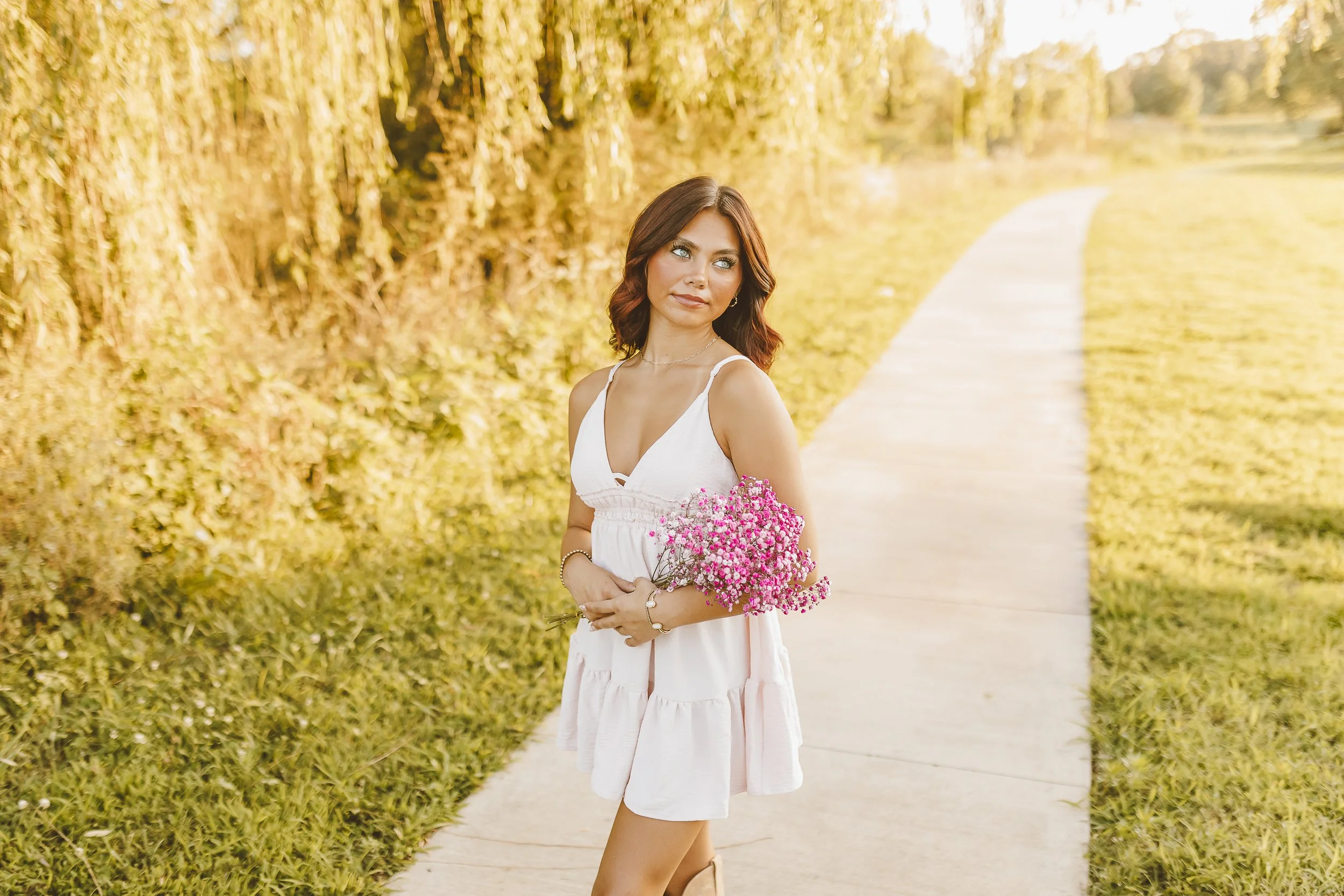 A young woman in a white dress holding a bouquet of pink flowers, standing on a pathway surrounded by yellow autumn foliage.