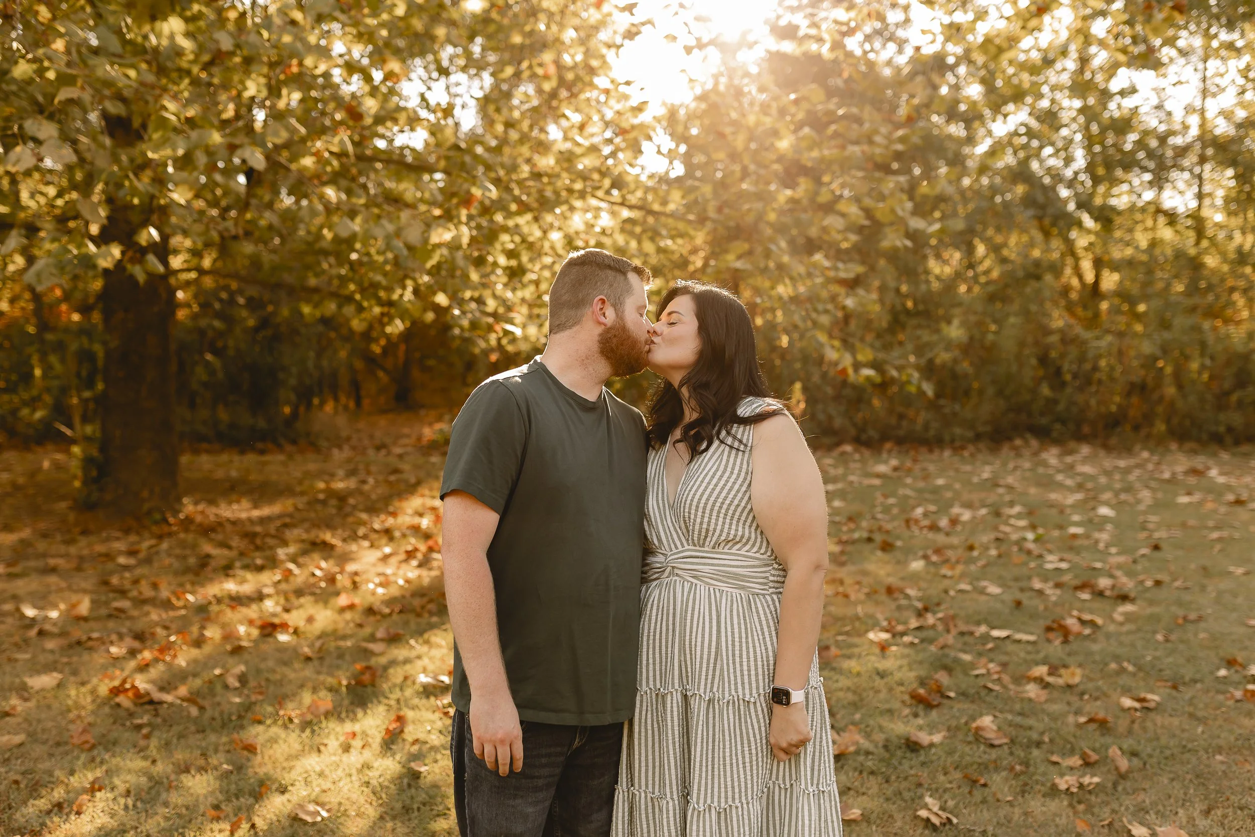 A couple is kissing outdoors during autumn, with golden sunlight filtering through trees with fall foliage.