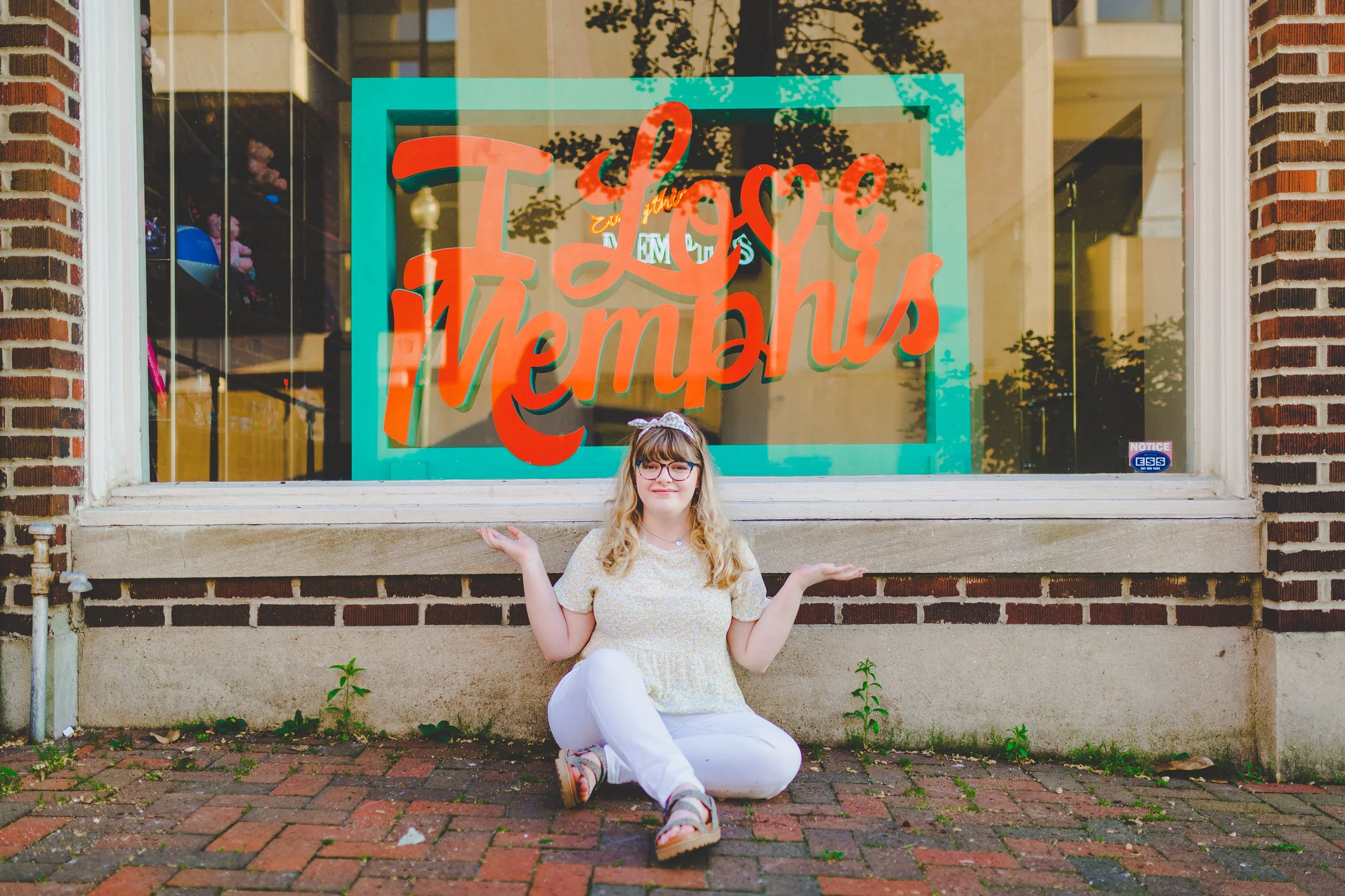 A young woman with glasses and curly hair sitting on brick sidewalk in front of a large window with a bright red and orange sign that says 'Love Memphis'. She is wearing a yellow shirt with white pants and has a pink headband. She is smiling with her arms raised and palms up.