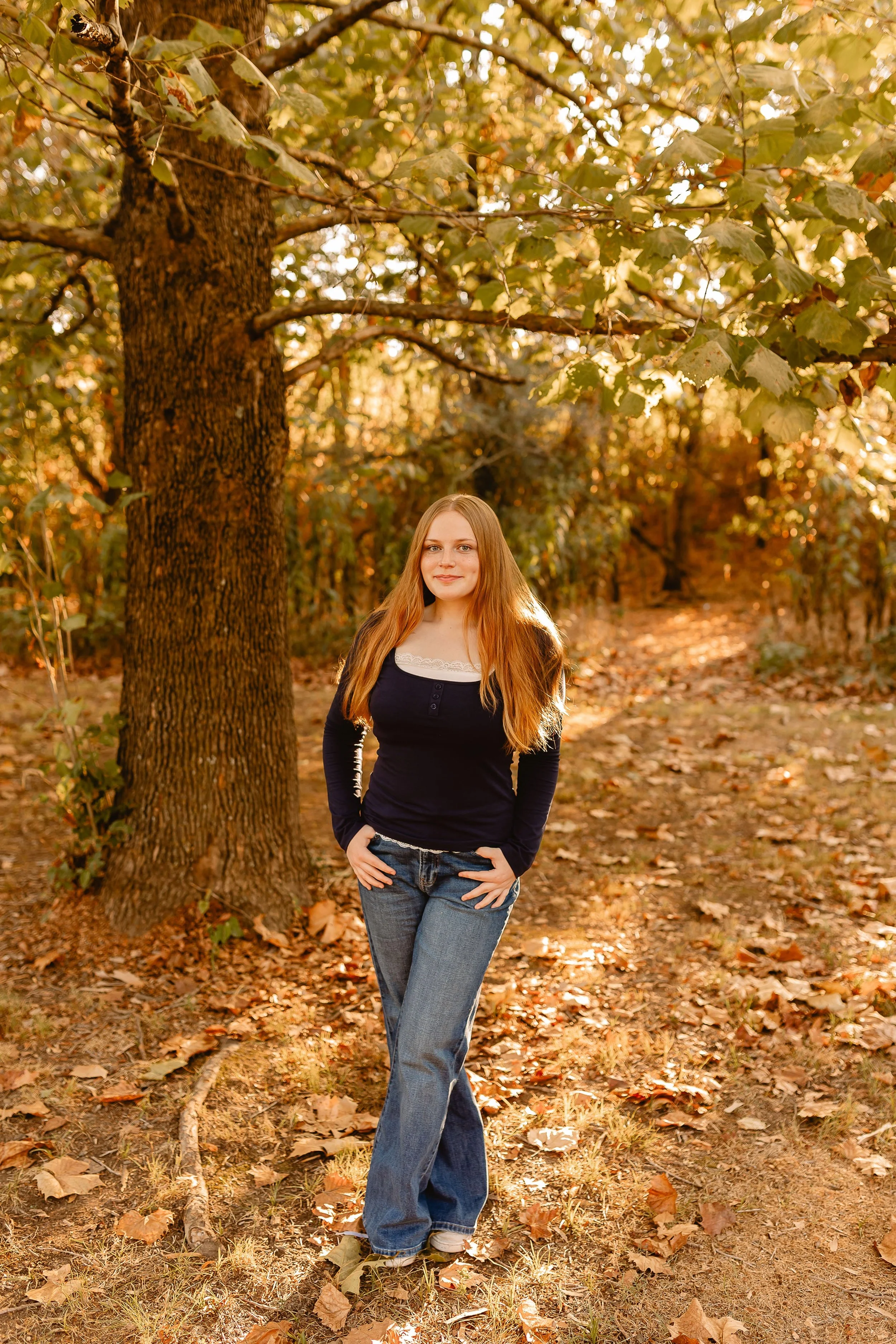 A young woman with long red hair standing on a leaf-covered ground in an autumn forest, wearing jeans and a black long sleeve shirt, with trees and foliage in the background.