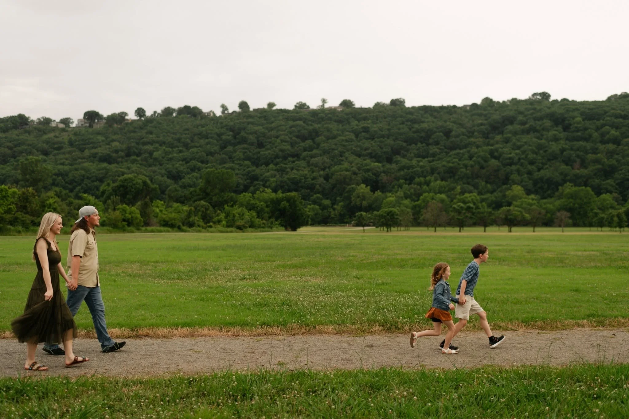 A family of five walking along a dirt path in a lush green park with trees and a hill in the background. The sky is overcast.