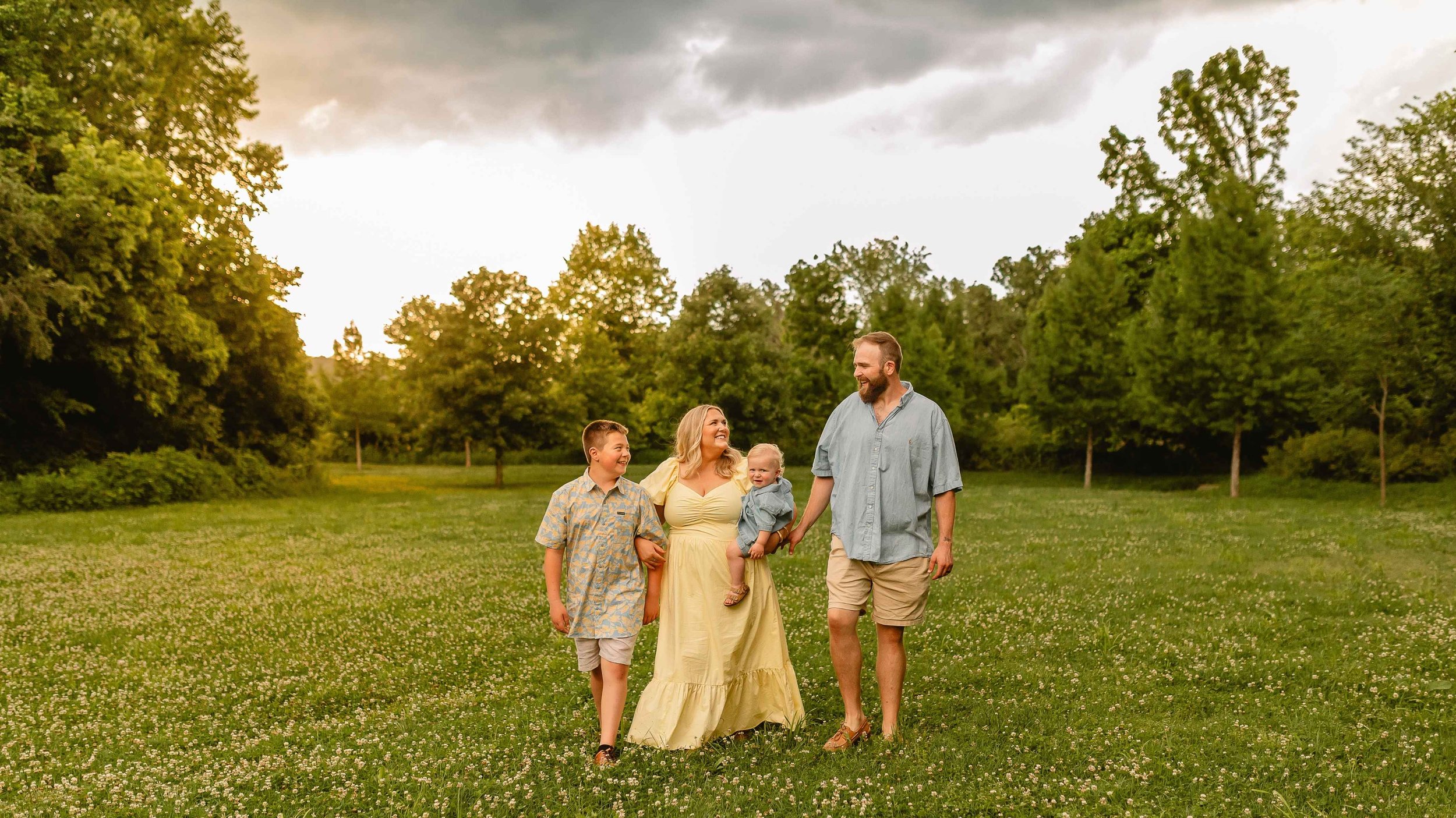 A family of four walking through a green meadow in the evening, with trees and a cloudy sky in the background.