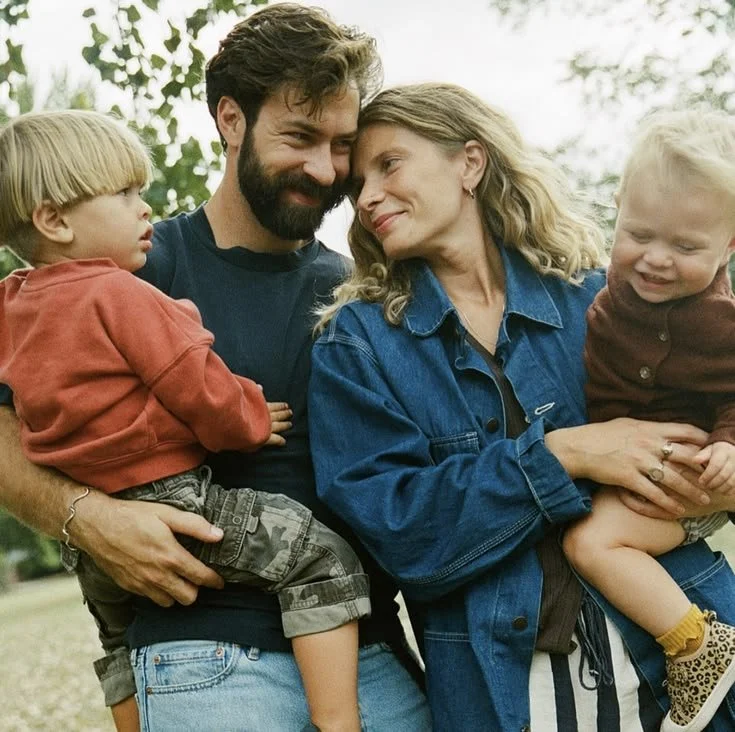 A family of four outdoors, embracing each other. The father holds a young boy in a red hoodie, while the mother holds a young girl in a brown shirt. All are smiling and looking at each other, with trees in the background.