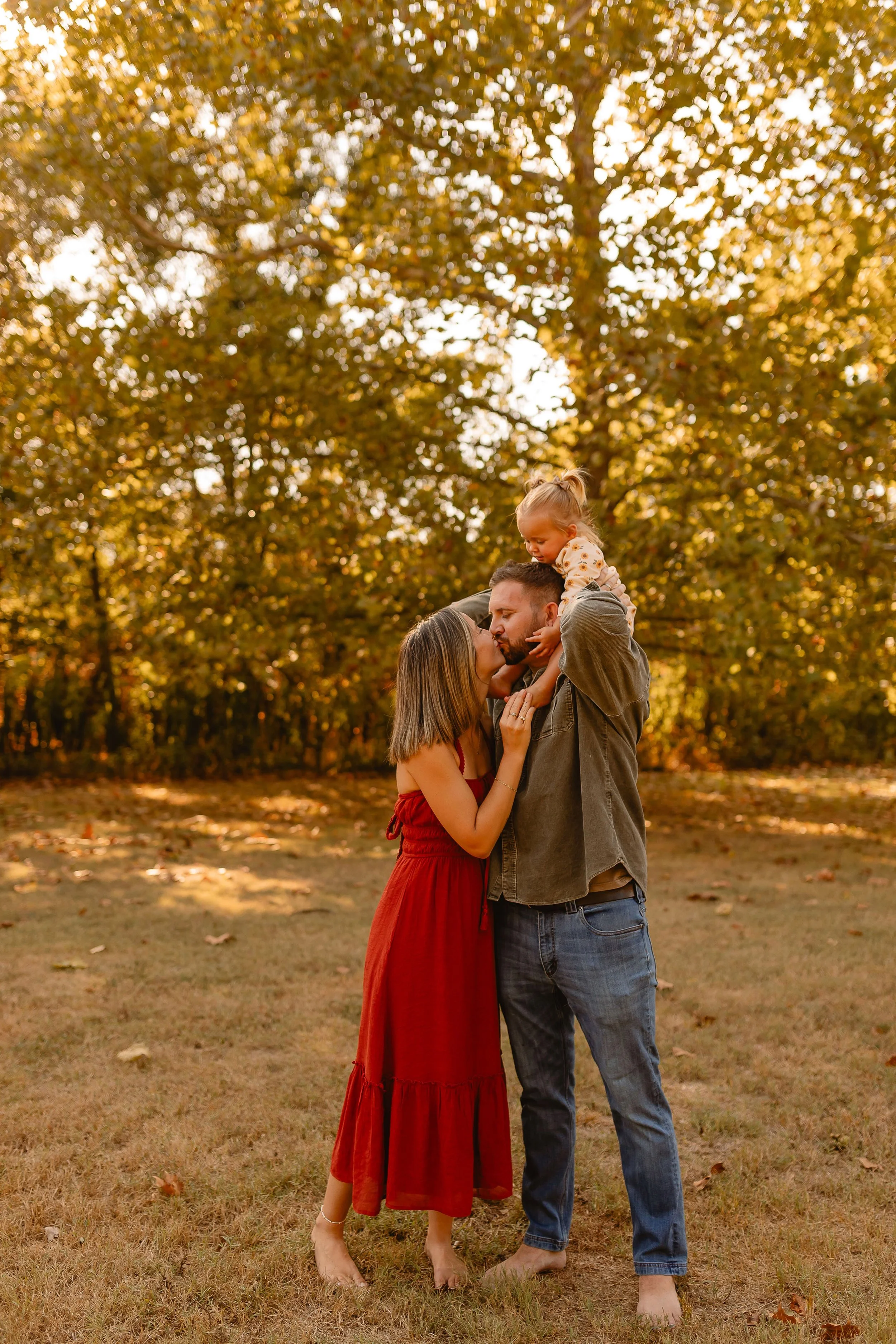 A family of three standing outdoors in a park during fall. The father, holding a young girl on his shoulders, is giving a kiss to the mother, who is wearing a red dress. The scene features trees with orange and yellow leaves.
