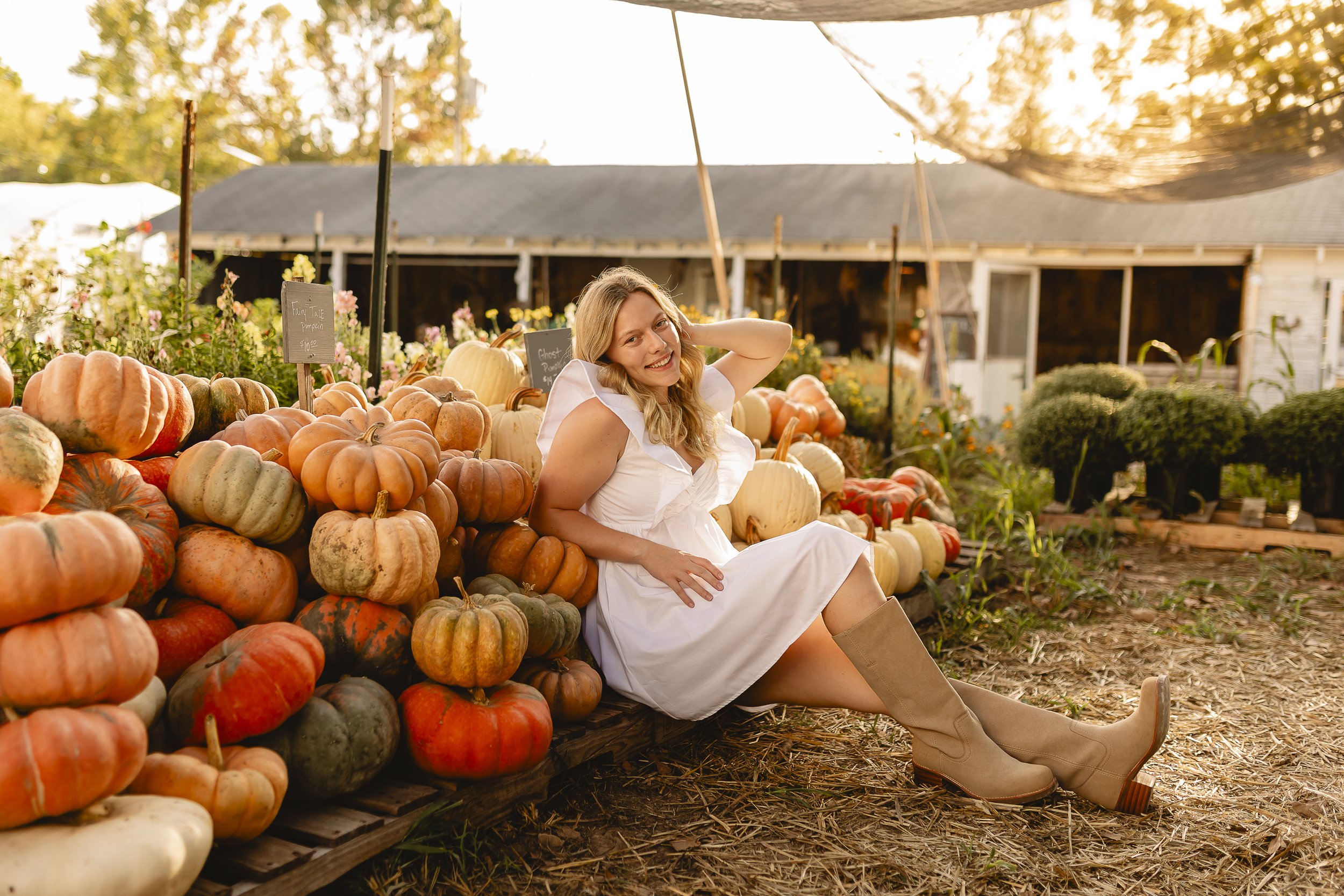 A woman in a white dress and tan boots sitting among pumpkins at a fall pumpkin patch or farm with a building and trees in the background during sunset.