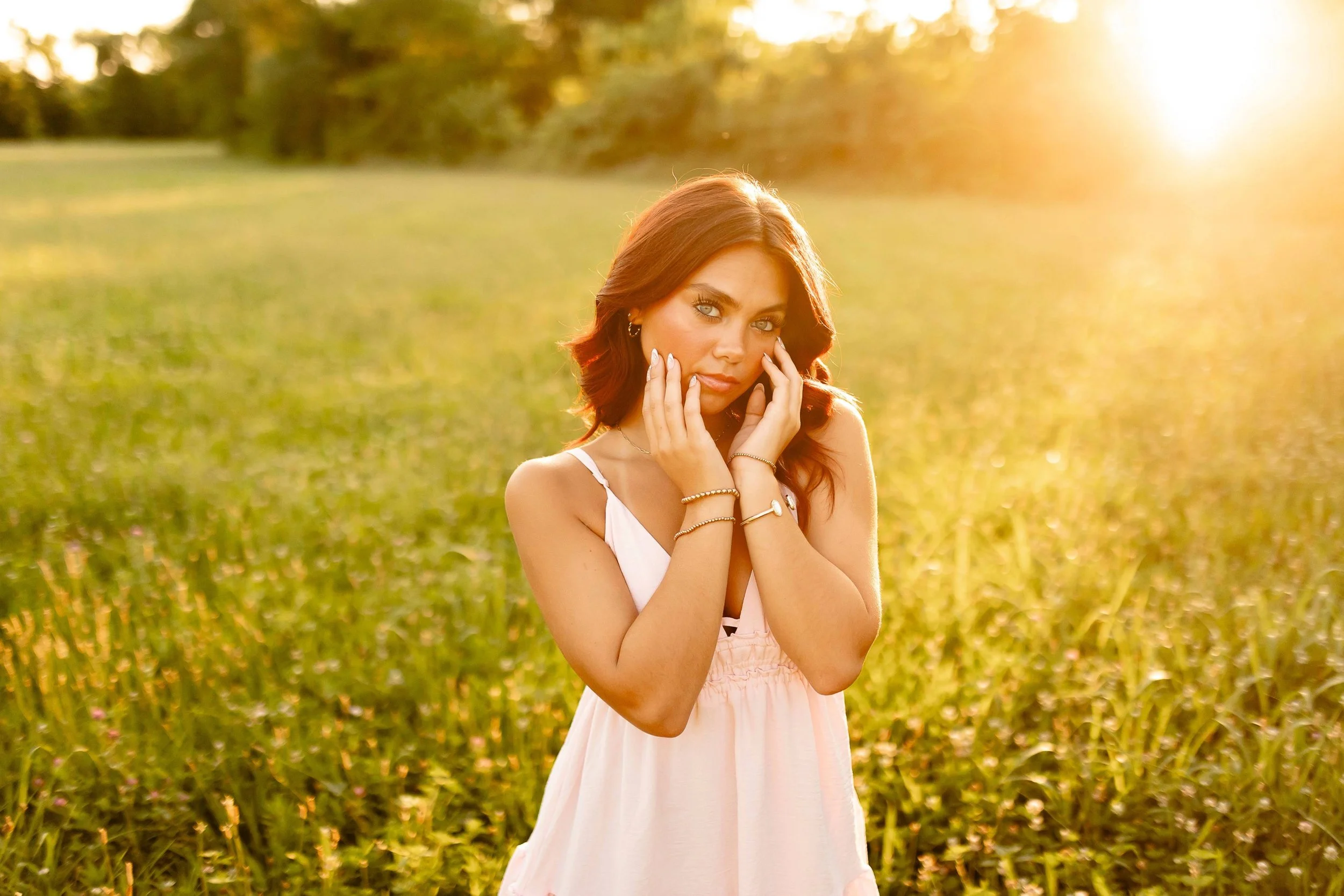 A young woman with red hair in a pink dress standing in a field at sunset, touching her face with both hands, looking at the camera.