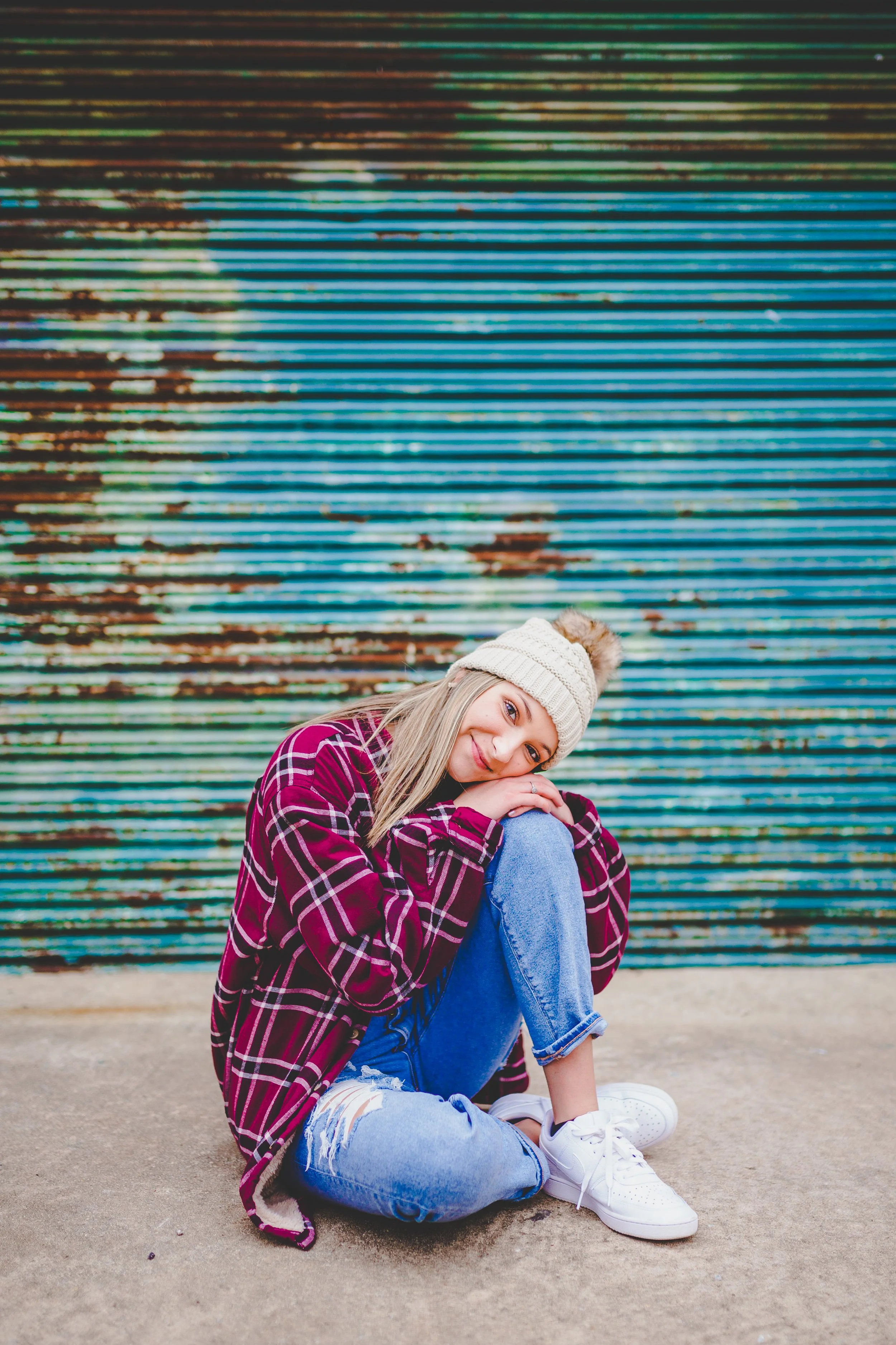 A smiling young woman with blonde hair sitting on concrete ground in front of a colorful, weathered metal wall. She is wearing a beige knit beanie, a maroon and black plaid shirt, ripped blue jeans, and white sneakers, with her arms wrapped around he