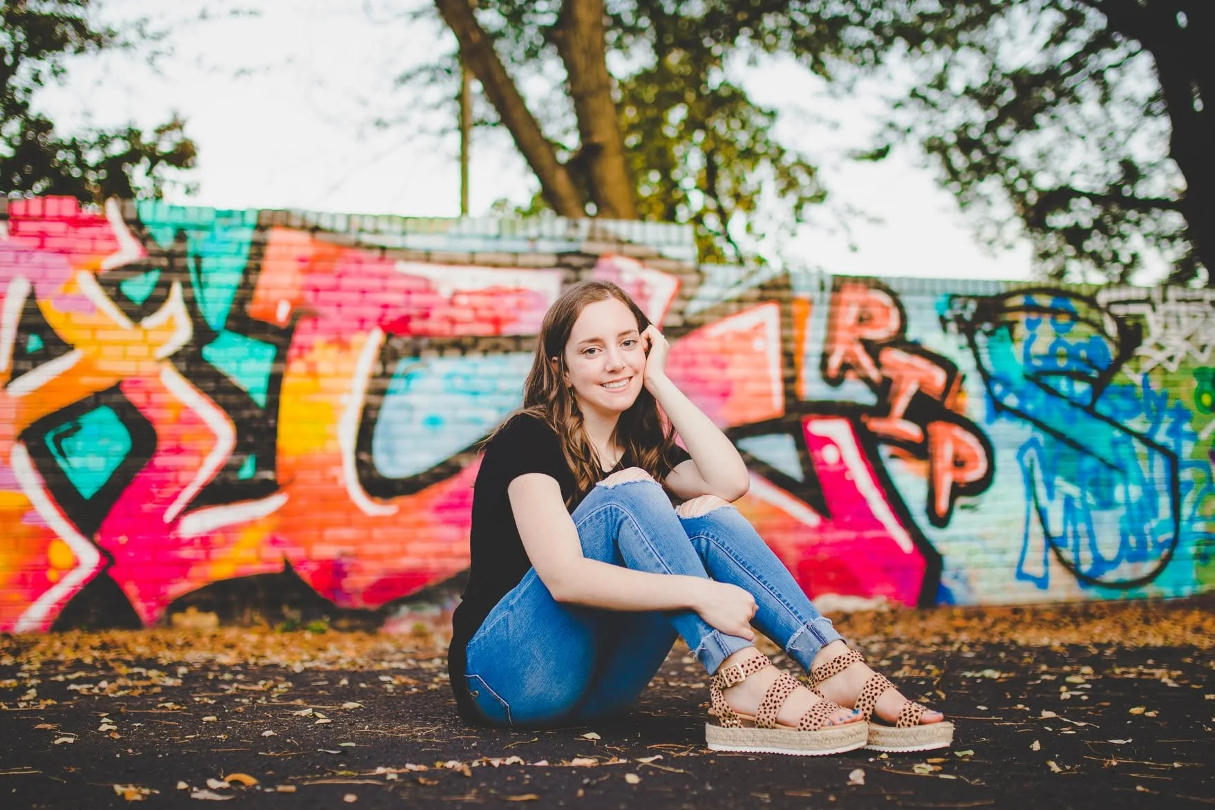 A young woman with long brown hair, wearing a black shirt, ripped jeans, and leopard print sandals, sitting on the ground in front of a colorful graffiti wall, with trees in the background.