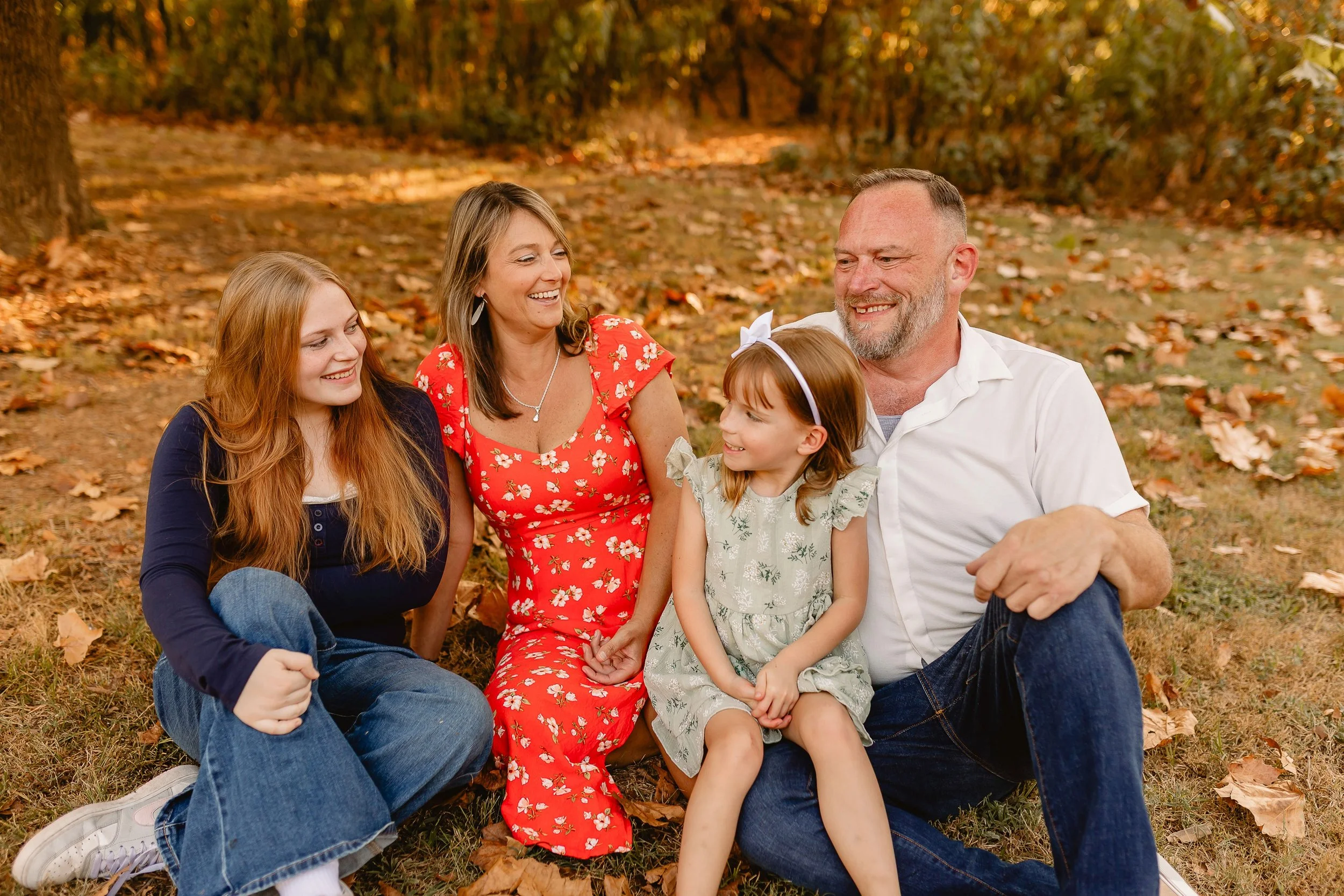 A family of five sitting on a grassy ground surrounded by fallen autumn leaves in a forest setting. The family includes a young woman with long red hair, an older woman with shoulder-length blonde hair, a young girl with a headband, a middle-aged man with short hair and beard, all smiling and enjoying each other's company.