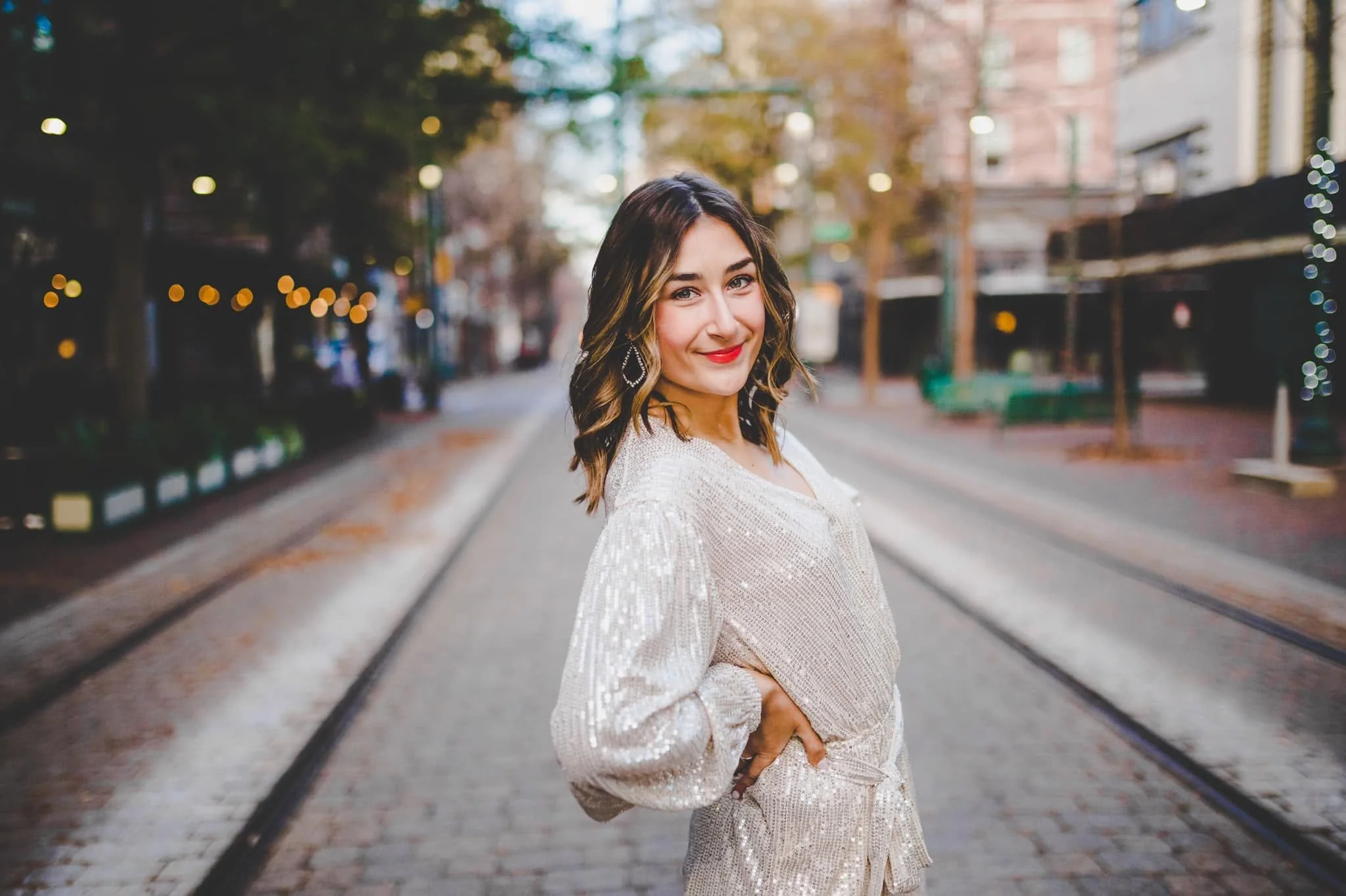 A woman with wavy dark hair and red lipstick standing on tram tracks in an urban area during fall, smiling and looking at the camera.