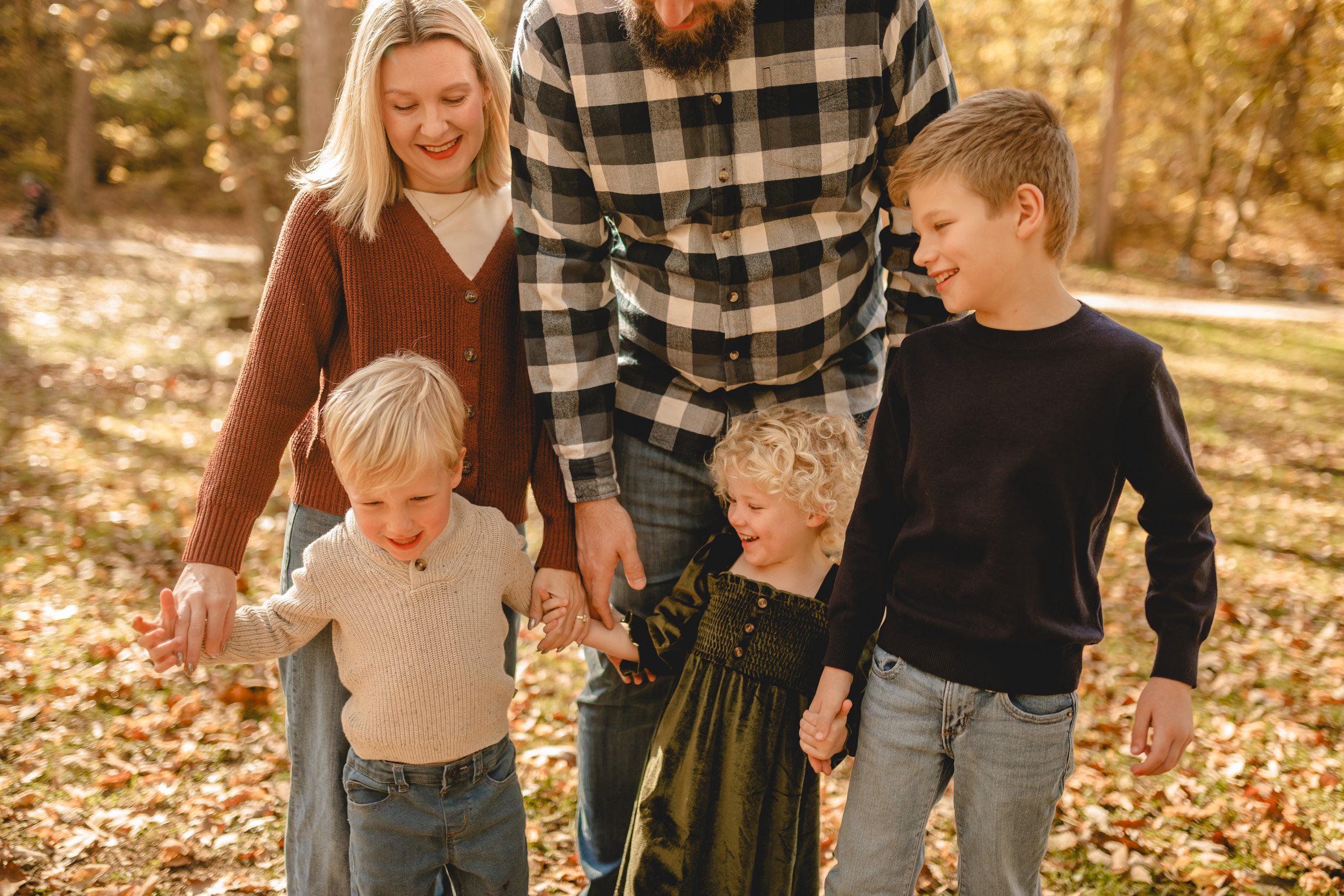 A family of six children and adults holding hands and smiling outdoors during fall, surrounded by autumn leaves.