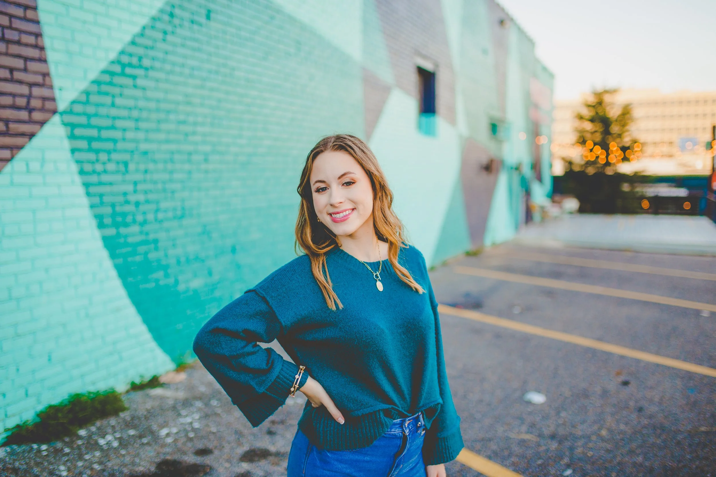 A young woman with light skin and wavy, shoulder-length hair smiling and standing outdoors in front of a colorful mural on a brick wall. She is wearing a blue sweater, jeans, and a necklace, with her hand on her hip. The background shows a parking lot and distant buildings with trees and warm lighting.