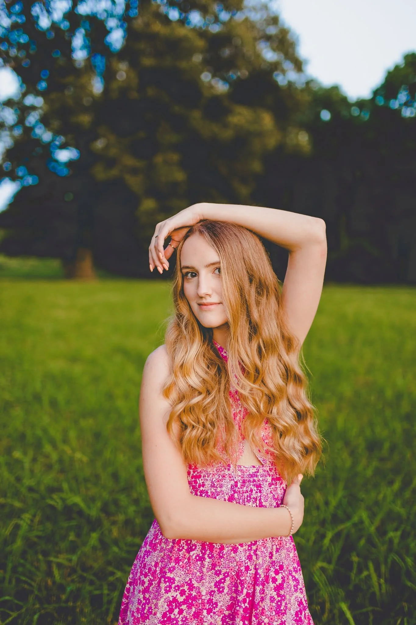A young woman with long, curly red hair wearing a pink floral dress with her right arm raised above her head, standing outdoors on a grassy field with trees in the background.