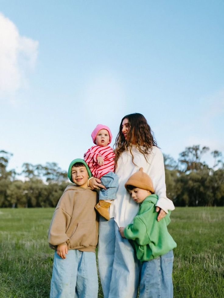 A woman with three children standing outdoors in a grassy field with trees in the background during daytime. The woman is holding a toddler, while two older children stand beside her.