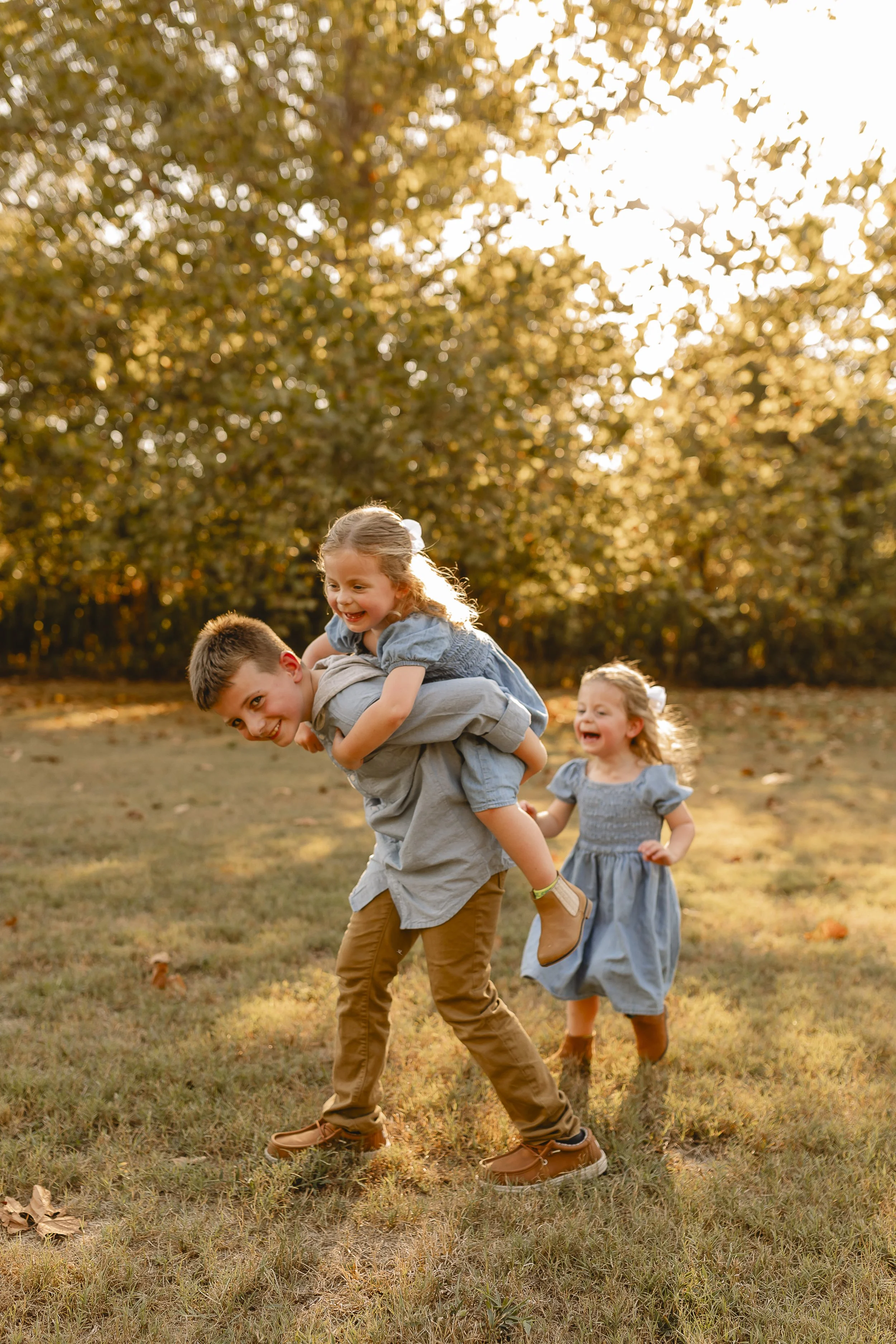 A group of children playing outdoors in a park during autumn, with one boy giving a piggyback ride to a girl, both smiling and enjoying themselves, with two other children following behind. The background features trees with orange leaves and sunligh