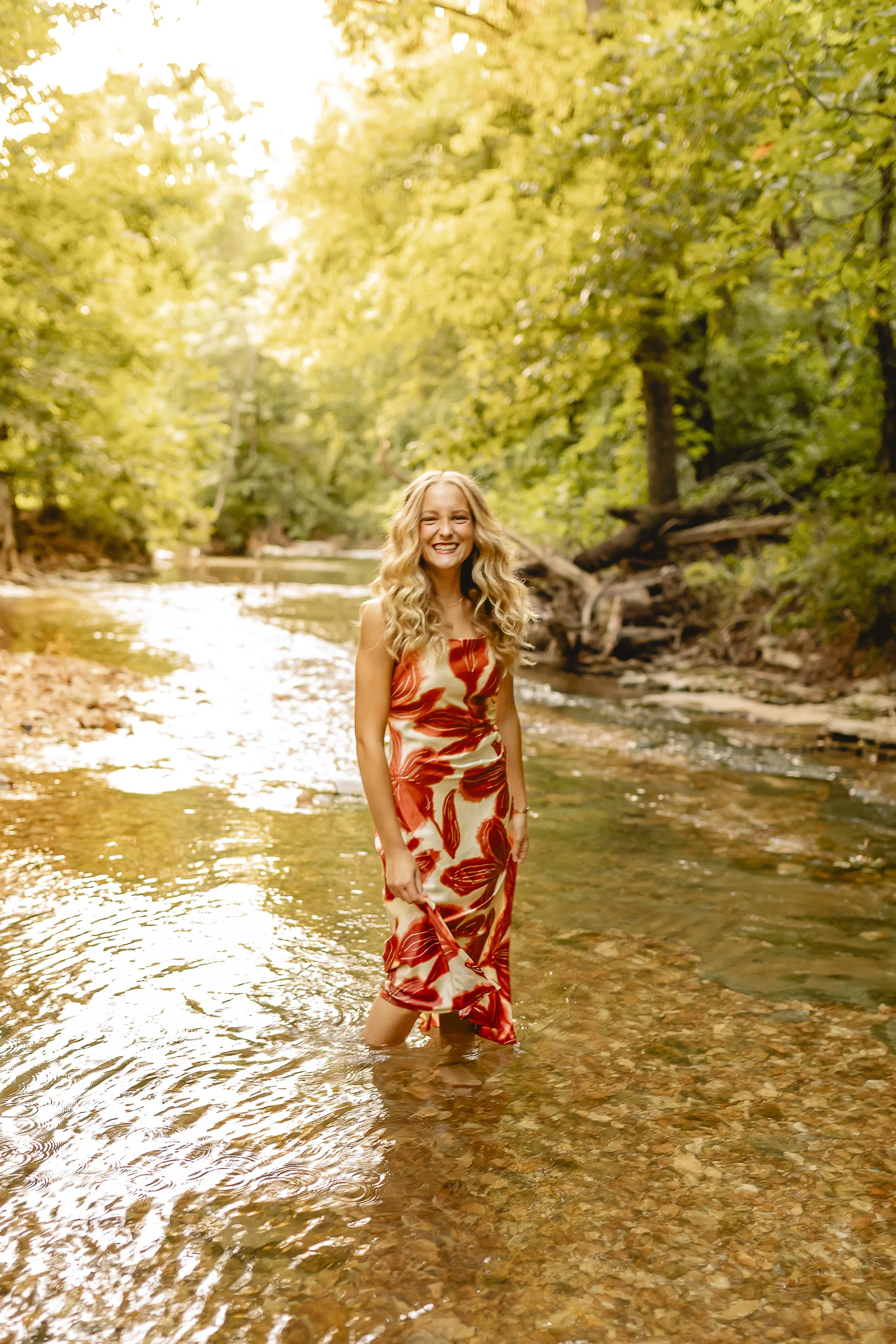 A smiling woman standing knee-deep in a creek surrounded by green trees, wearing a colorful dress with red, orange, and beige patterns.